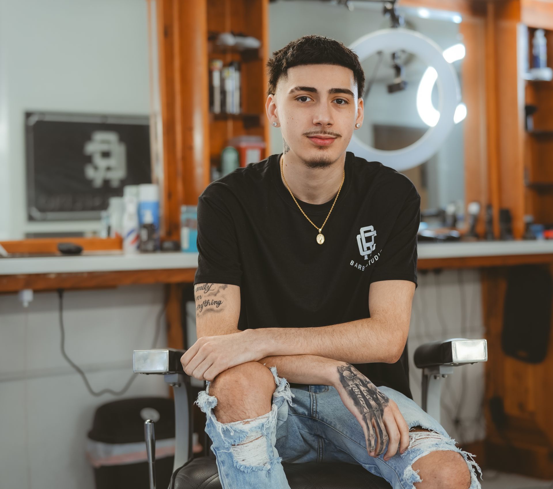 Man with a goatee and ripped jeans sits in a barber chair. He's wearing a black shirt, a gold chain, and has arm tattoos.