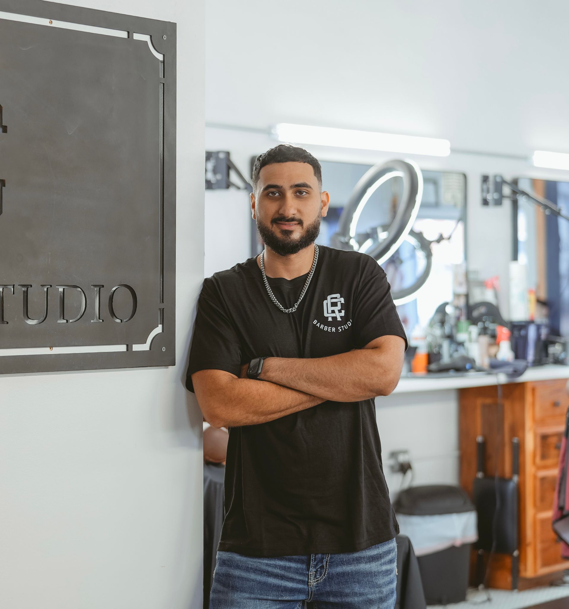 Man in black t-shirt, arms crossed, leans against a sign in a barber shop.