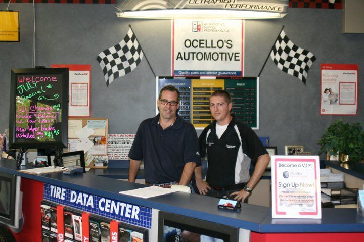 Two service workers stand behind the counter at Ocello's Automotive, beneath checkered racing flags. | Ocello’s Automotive Center