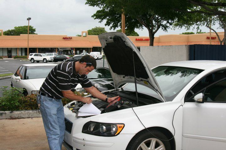 A person wearing a striped shirt examines the engine of a white car parked in a lot with a strip mall in the background. | Ocello’s Automotive Center