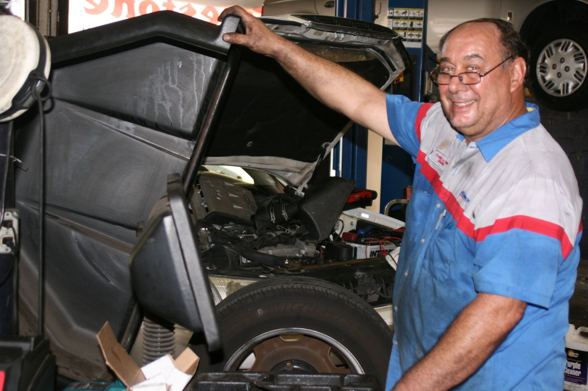 A smiling mechanic in a blue and white uniform stands in an auto shop with the hood of a vehicle open. | Ocello’s Automotive Center