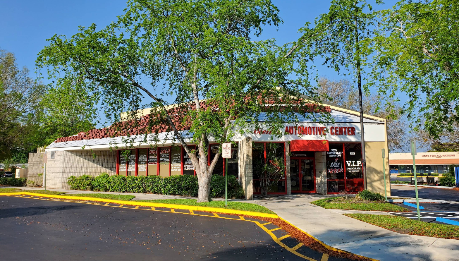 A one-story commercial building with red-tiled roofing, glass storefront windows, and landscaping under a clear blue sky. | Ocello’s Automotive Center