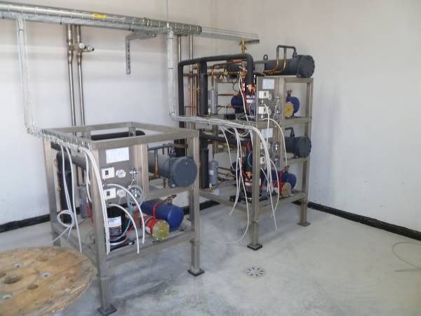 Attrezzature industriali in acciaio inossidabile in una stanza dalle pareti bianche, con tubi, pompe e manometri.