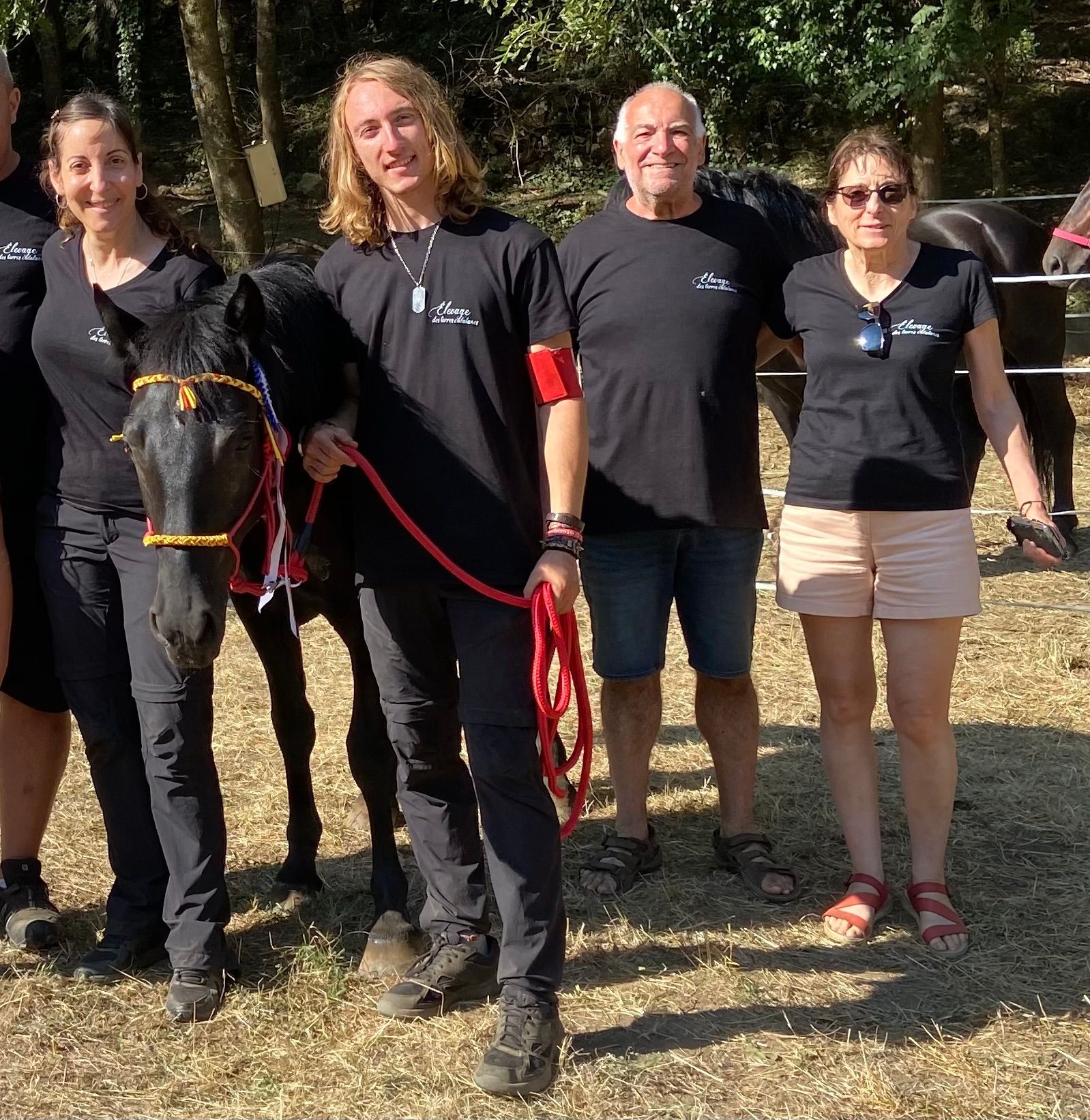 Un groupe pose avec un cheval noir. Les personnes portent des chemises noires ; le cheval arbore une coiffe décorative. En extérieur, il fait beau.