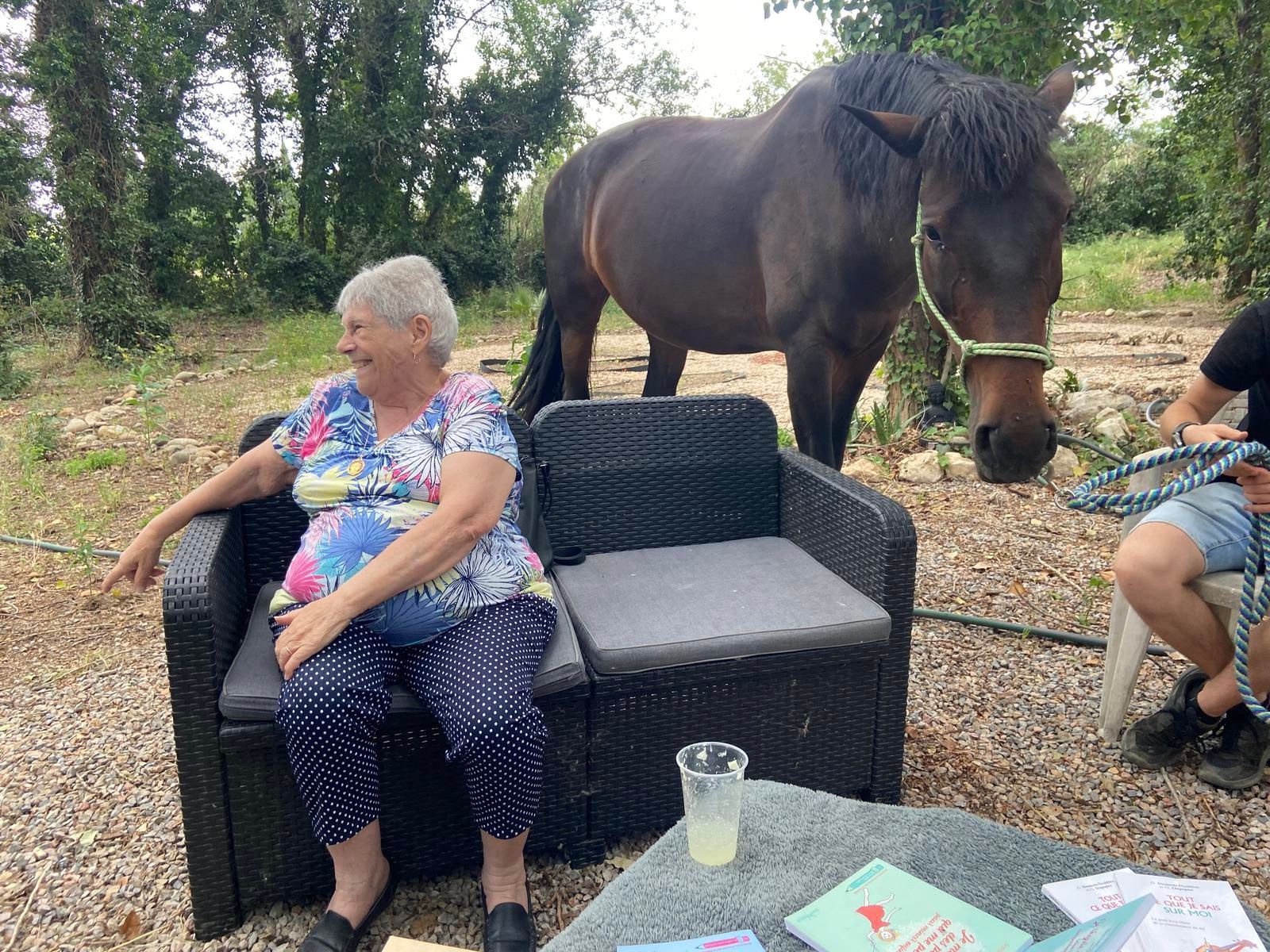 Une femme est assise sur un canapé en osier, un cheval à proximité, un homme tenant une corde. Extérieur, sur un sol gravillonné et verdoyant.