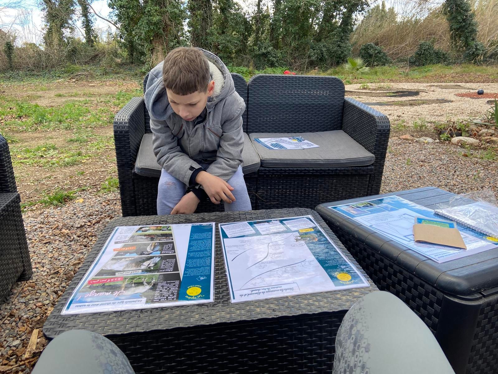 Une personne assise à une table en terrasse consulte des documents. Une deuxième personne est assise derrière la table.
