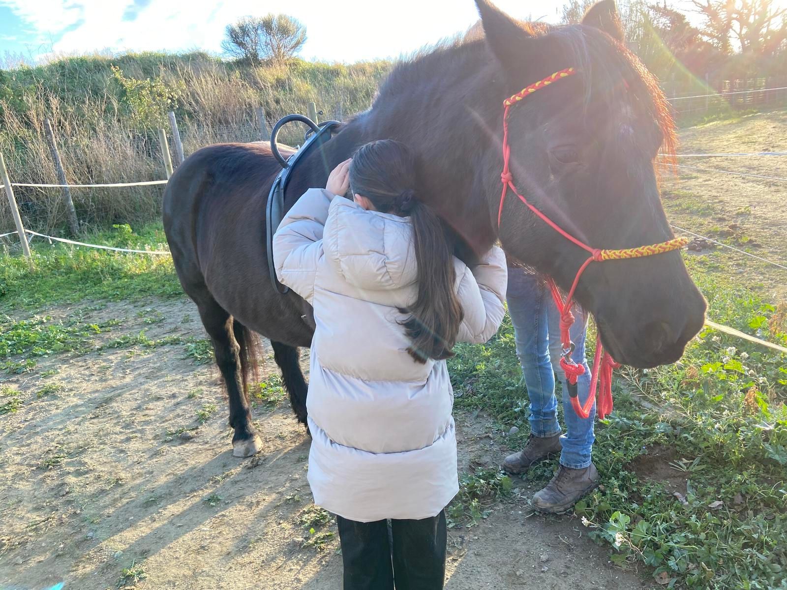Une personne vêtue d'une doudoune s'appuie contre un cheval sombre sellé, dans un décor herbeux en extérieur.