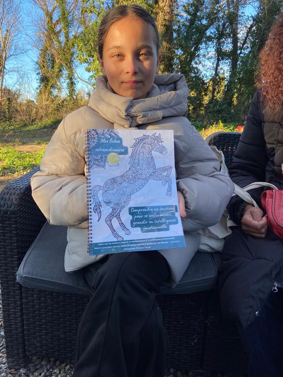 Une personne vêtue d'une doudoune tient à l'extérieur un livre à spirale, à la couverture bleue et blanche illustrée d'un cheval.