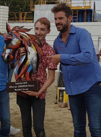 Un jeune homme et un homme tiennent un trophée en forme de cheval, en extérieur. Le jeune homme porte une chemise rouge et l'homme, une chemise bleue.