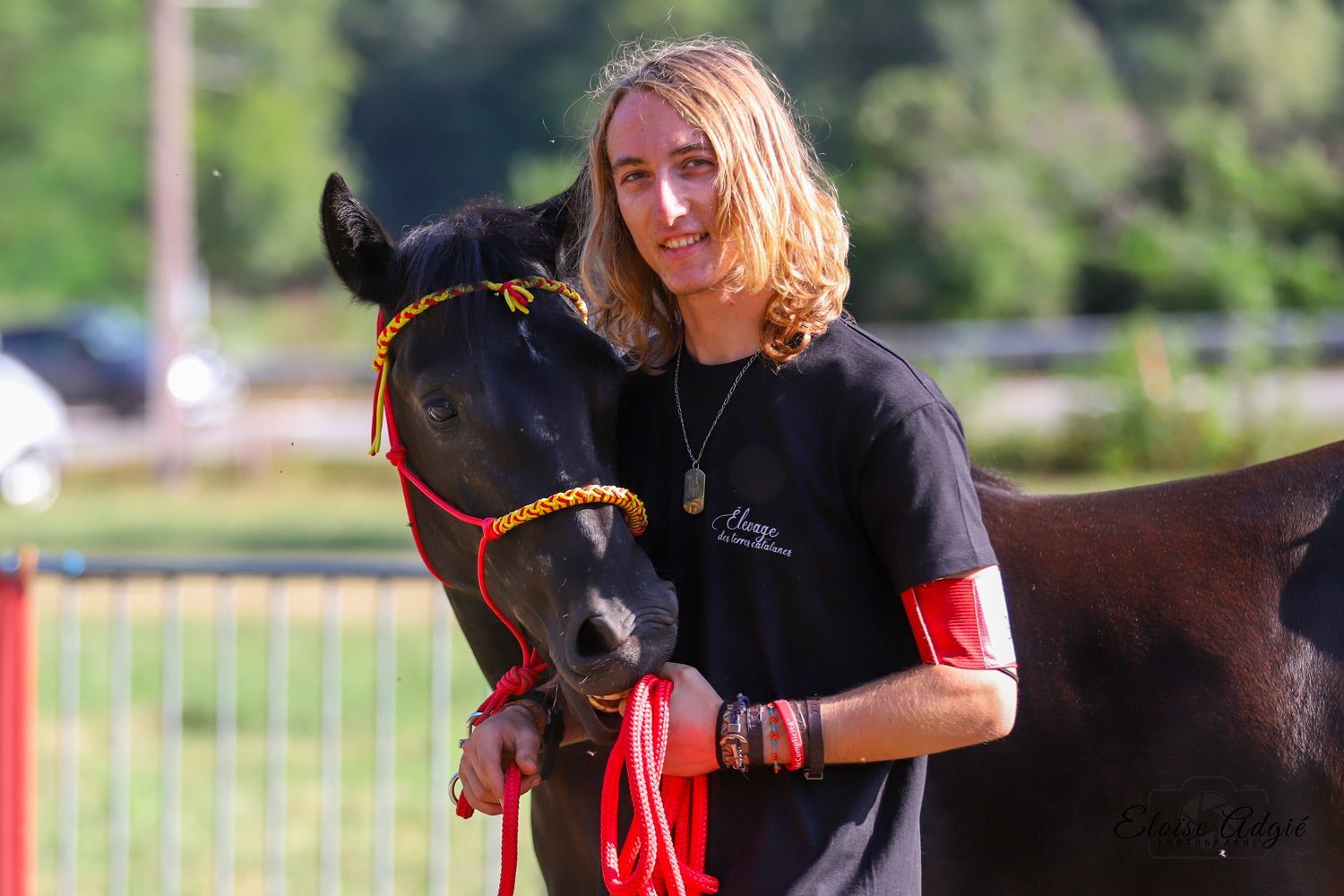 Un homme aux longs cheveux blonds tient un cheval noir portant un licol rouge et or ; en extérieur.
