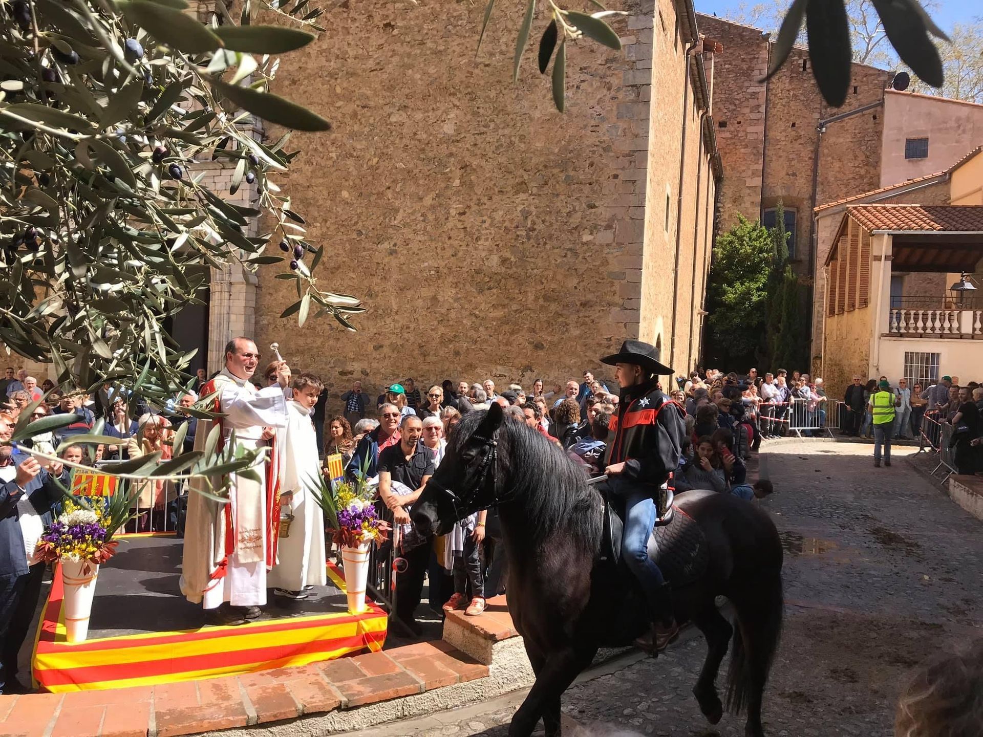 Une personne à cheval noir participe à une procession près d'une église. Un prêtre bénit les fidèles avec des fleurs sous le regard de la foule.