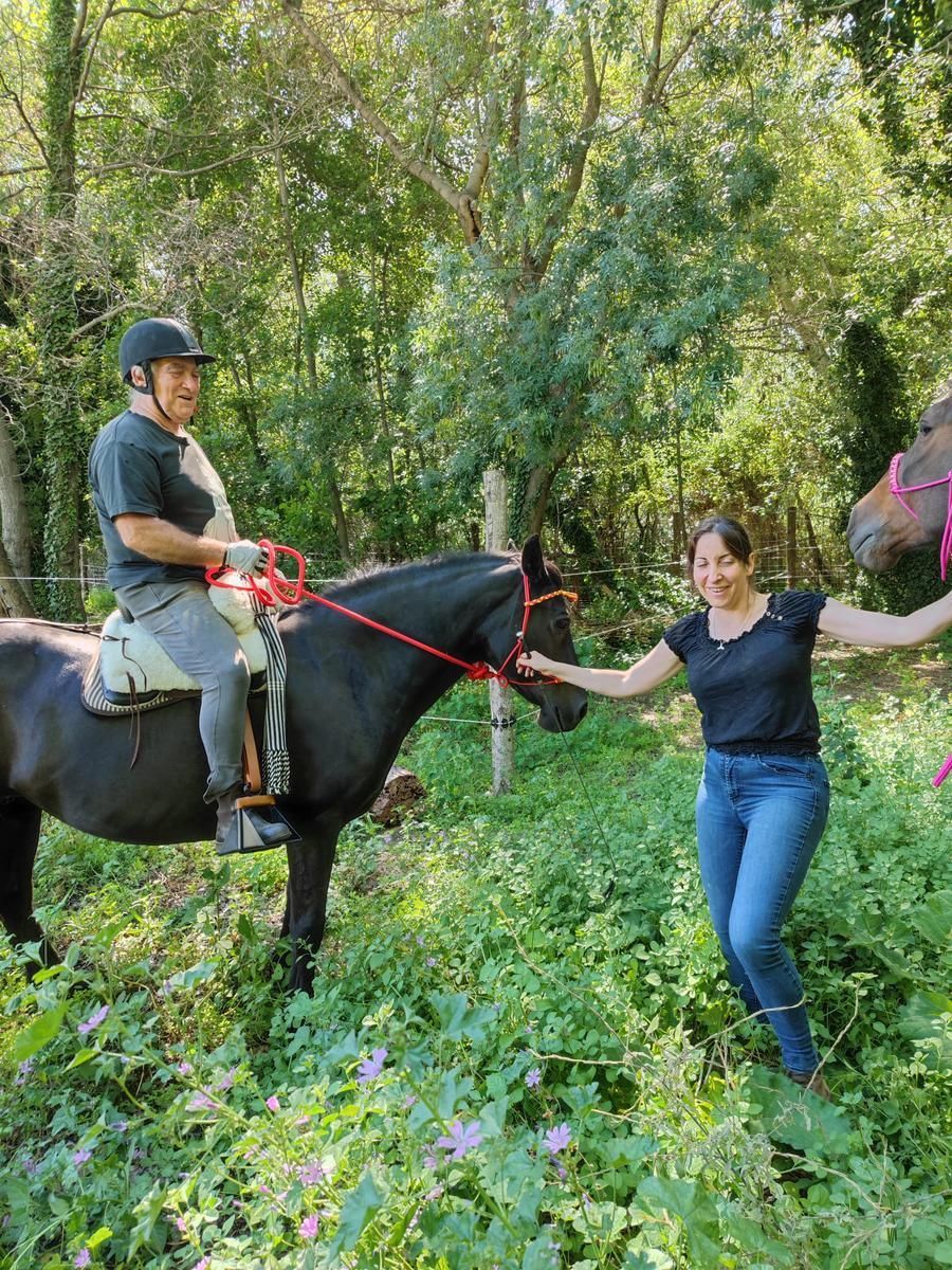 Une personne à cheval (un cheval noir), une autre femme tenant les rênes dans une zone boisée.