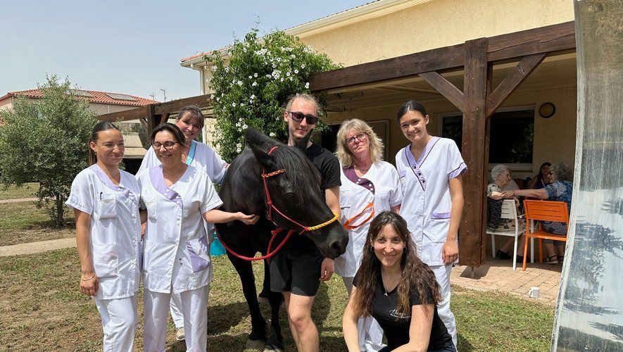 Un groupe pose avec un cheval noir à l'extérieur, près d'un bâtiment ; journée ensoleillée.