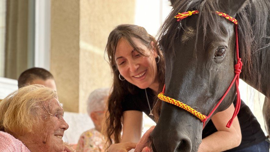 Une femme à cheval interagit avec une personne âgée en extérieur ; elle sourit et touche le cheval.