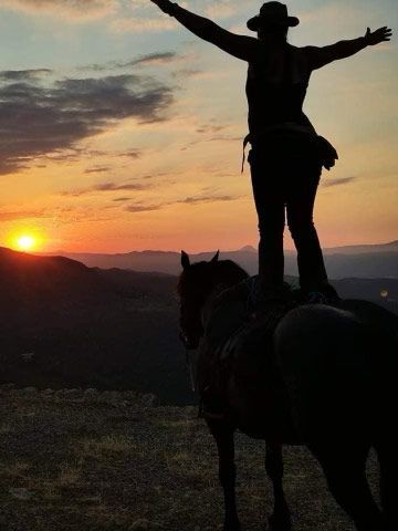 Silhouette d'une personne debout à cheval, les bras levés, se détachant sur un coucher de soleil sur les montagnes.