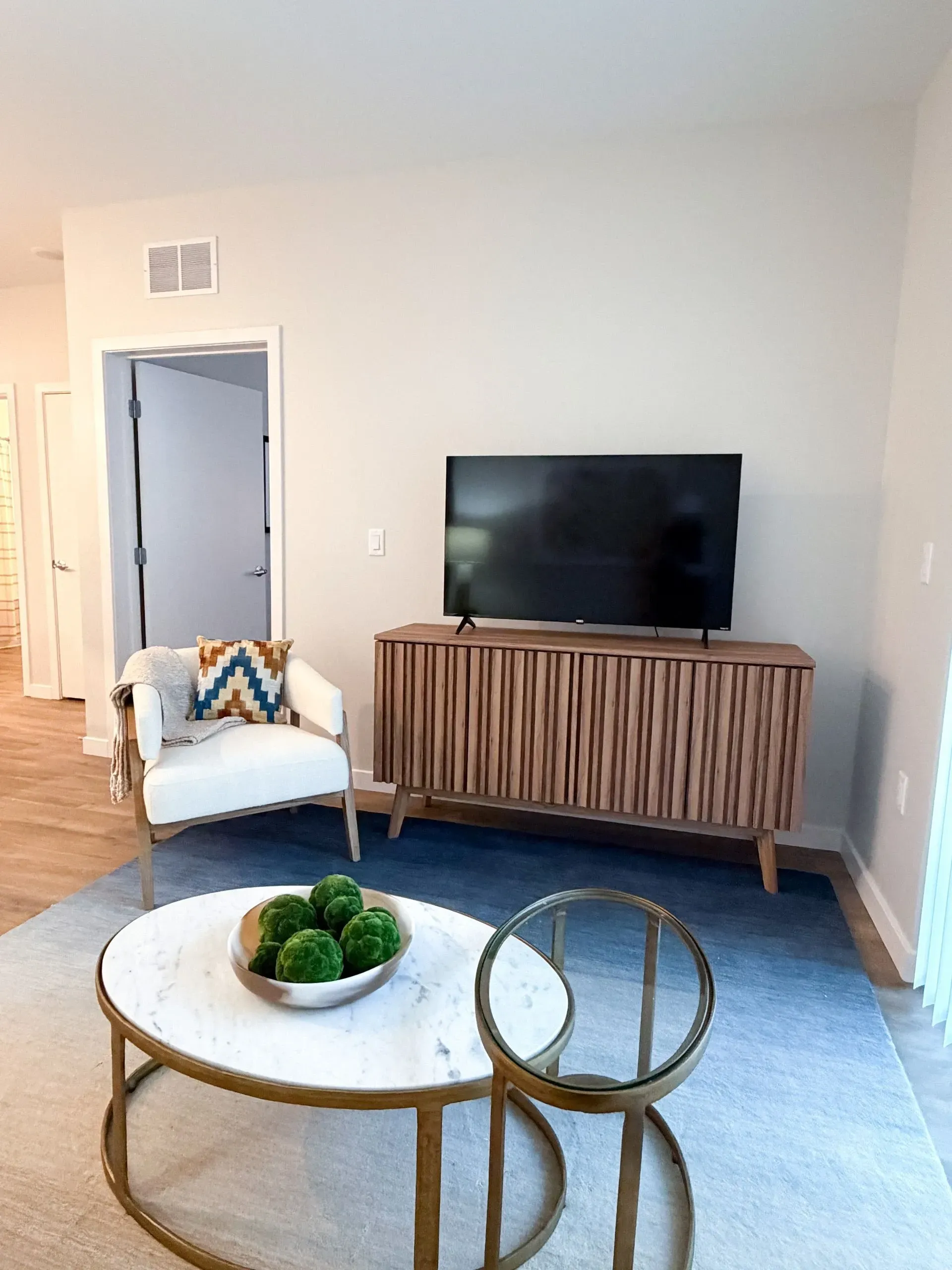 Living room with a flat-screen TV on a wooden slatted media console, white armchair, and round marble coffee table.