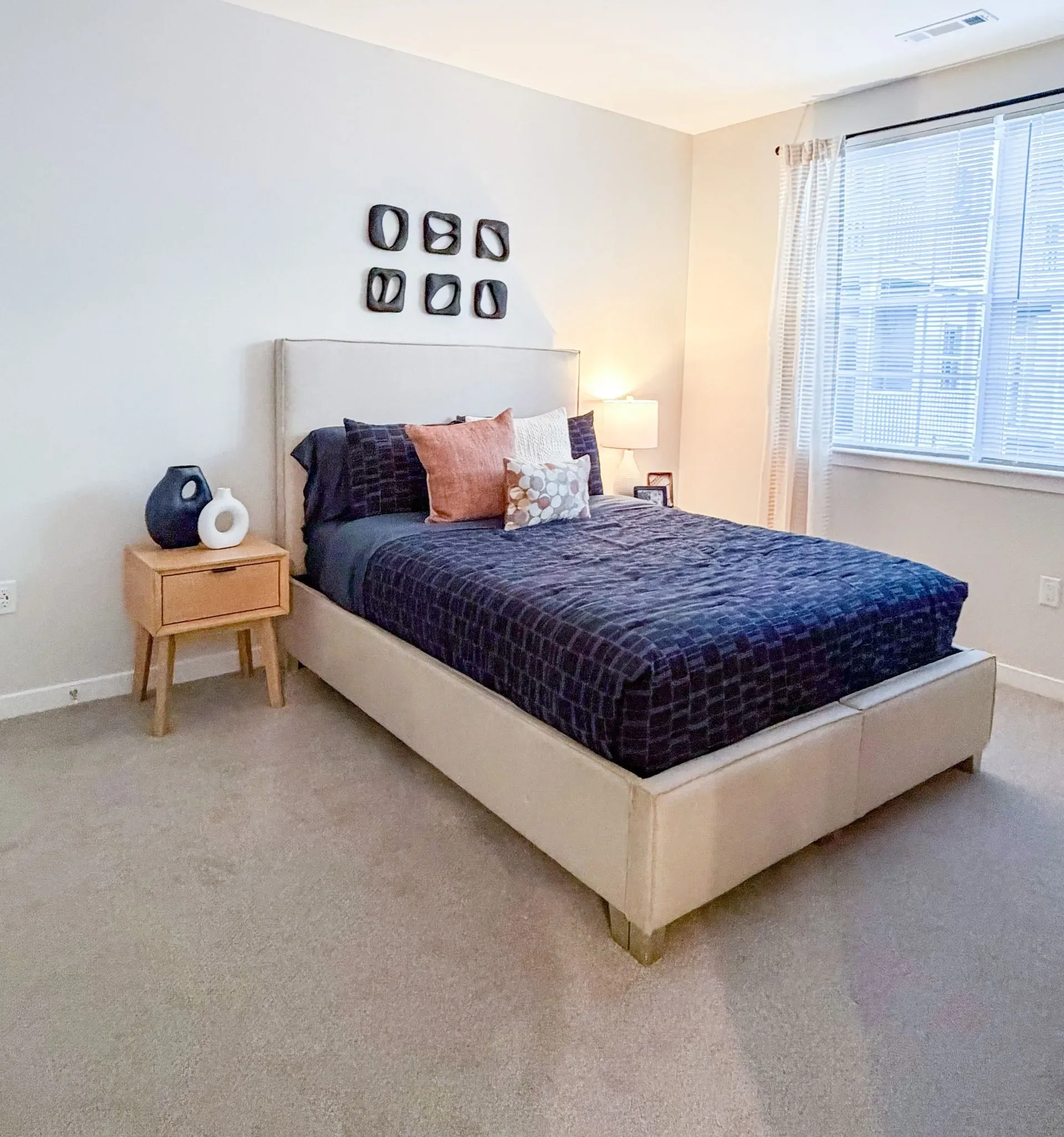 Bedroom with beige upholstered bed, nightstand, lamp, and window blinds.
