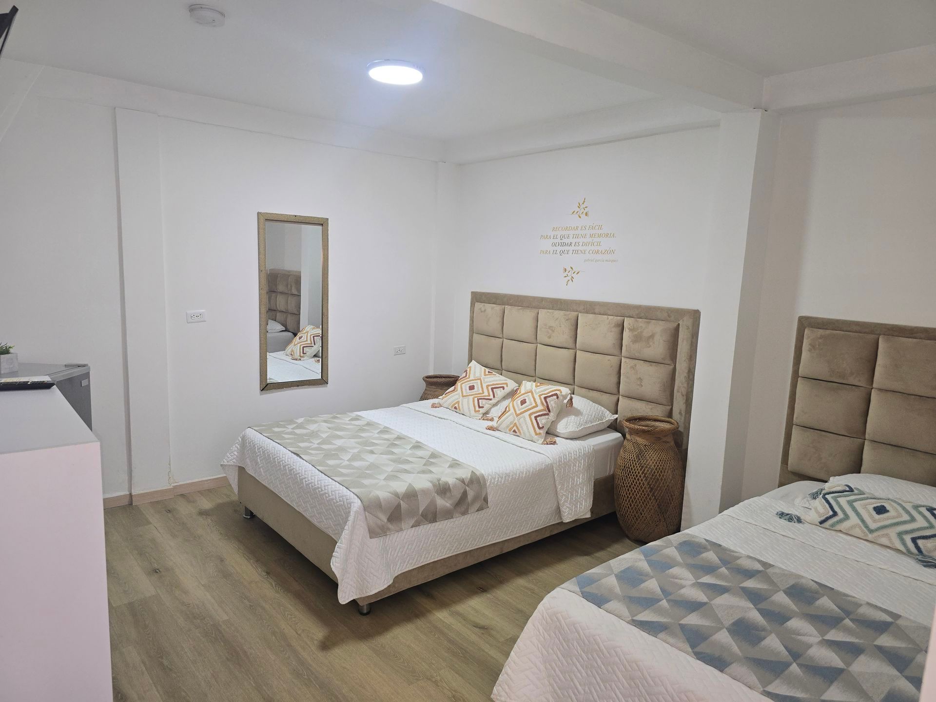 Una habitación de hotel con una cama de matrimonio y una cama individual, ambas con cabeceros acolchados de color beige, suelo de madera clara y un espejo.