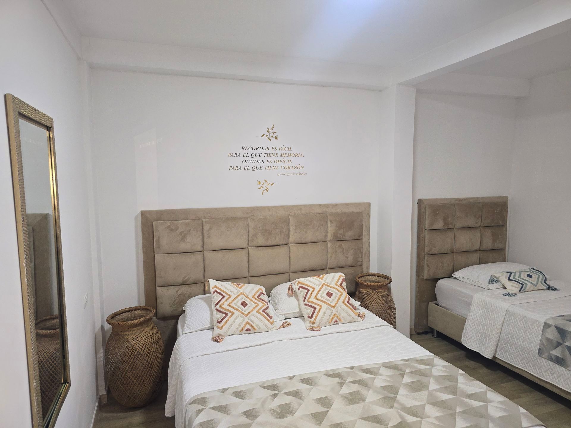 Un dormitorio con una cama doble y una cama individual con cabeceros acolchados de color beige, ropa de cama blanca y almohadas estampadas.