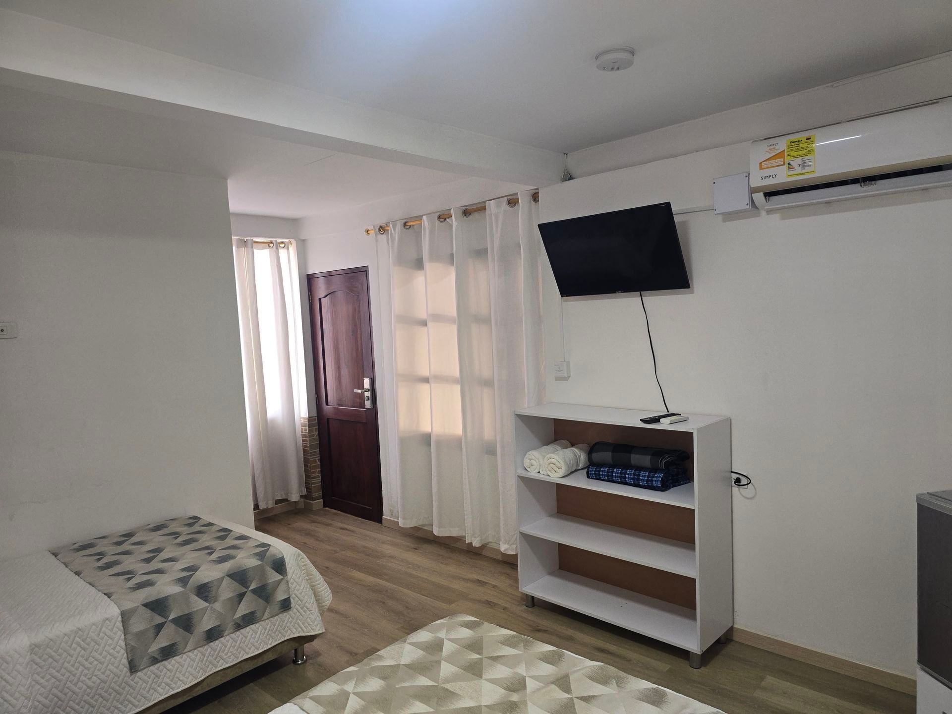 Un dormitorio con dos camas, una estantería blanca para toallas, un televisor montado en la pared y un aparato de aire acondicionado blanco.