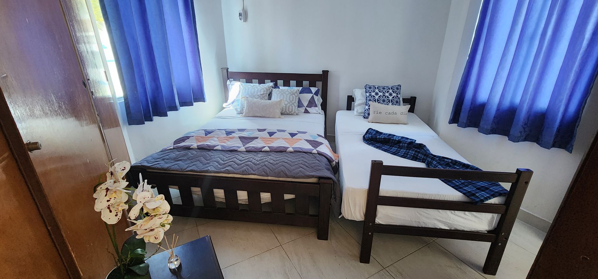 Un dormitorio con una cama de matrimonio y una cama individual, ambas con cortinas y ropa de cama azules, sobre un suelo de baldosas.