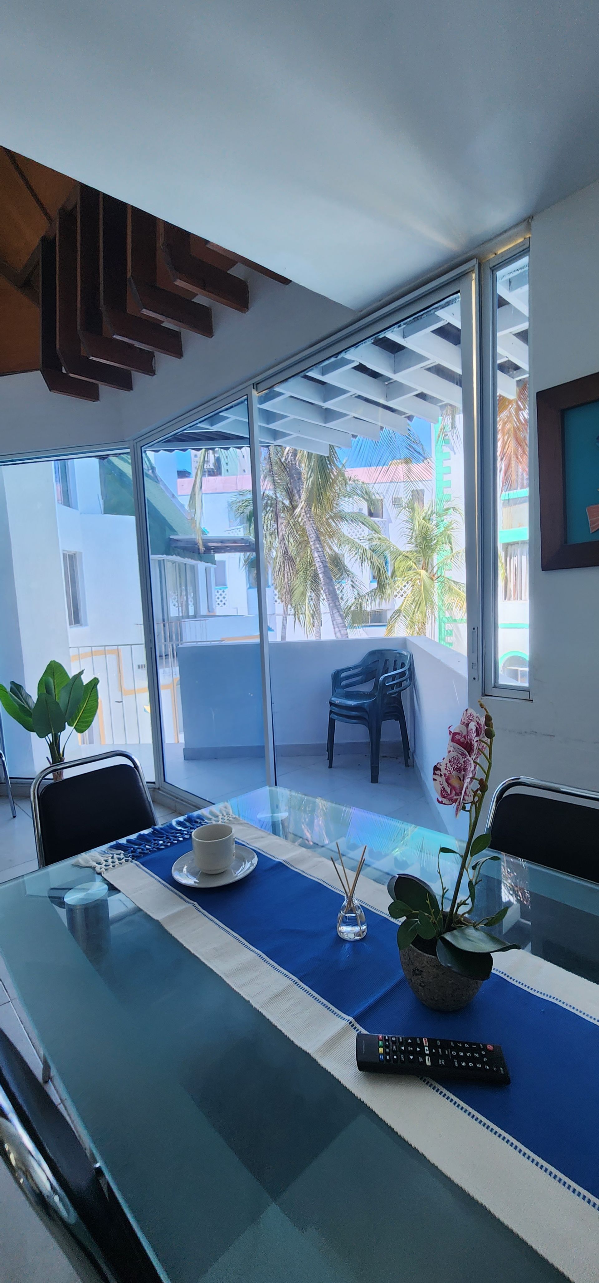 Una mesa de comedor con un camino de mesa azul y una pequeña planta, colocada frente a una gran ventana con vistas a un balcón soleado.