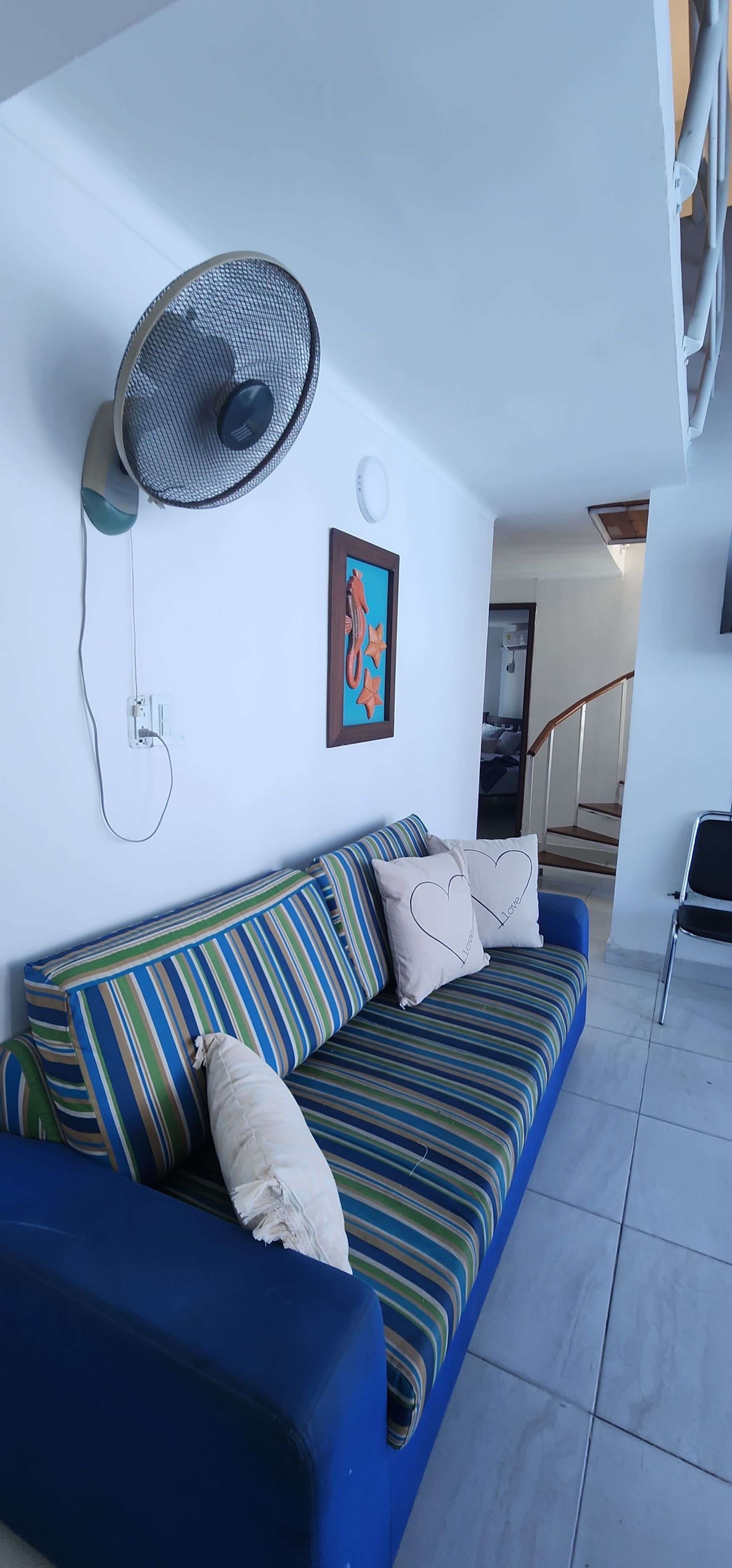 Un sofá con tapicería de rayas azules y verdes y dos cojines blancos, situado debajo de un ventilador de pared en una habitación luminosa.