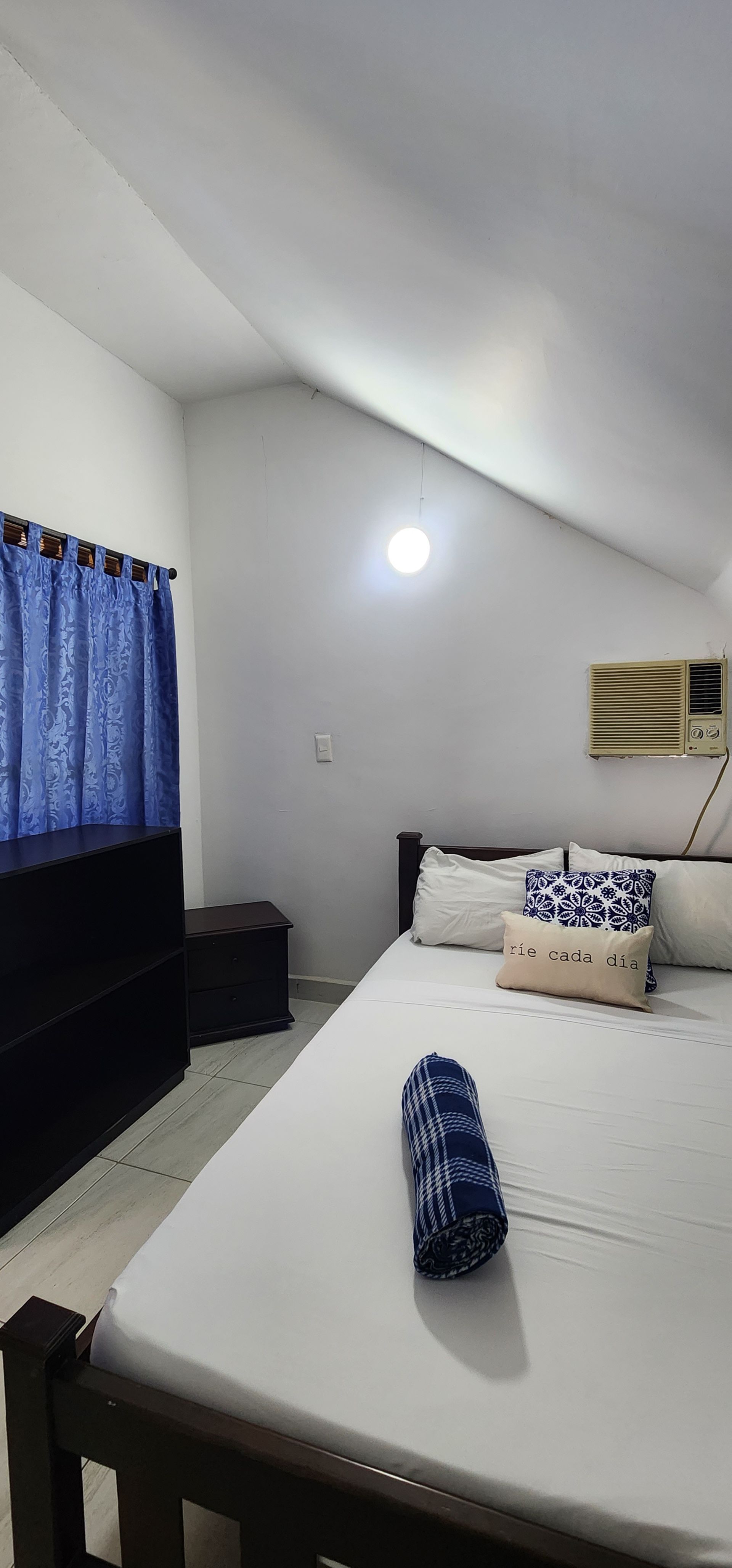 Un dormitorio con techo blanco inclinado, una cama blanca con almohadas estampadas, cortinas azules y una cómoda de madera negra.