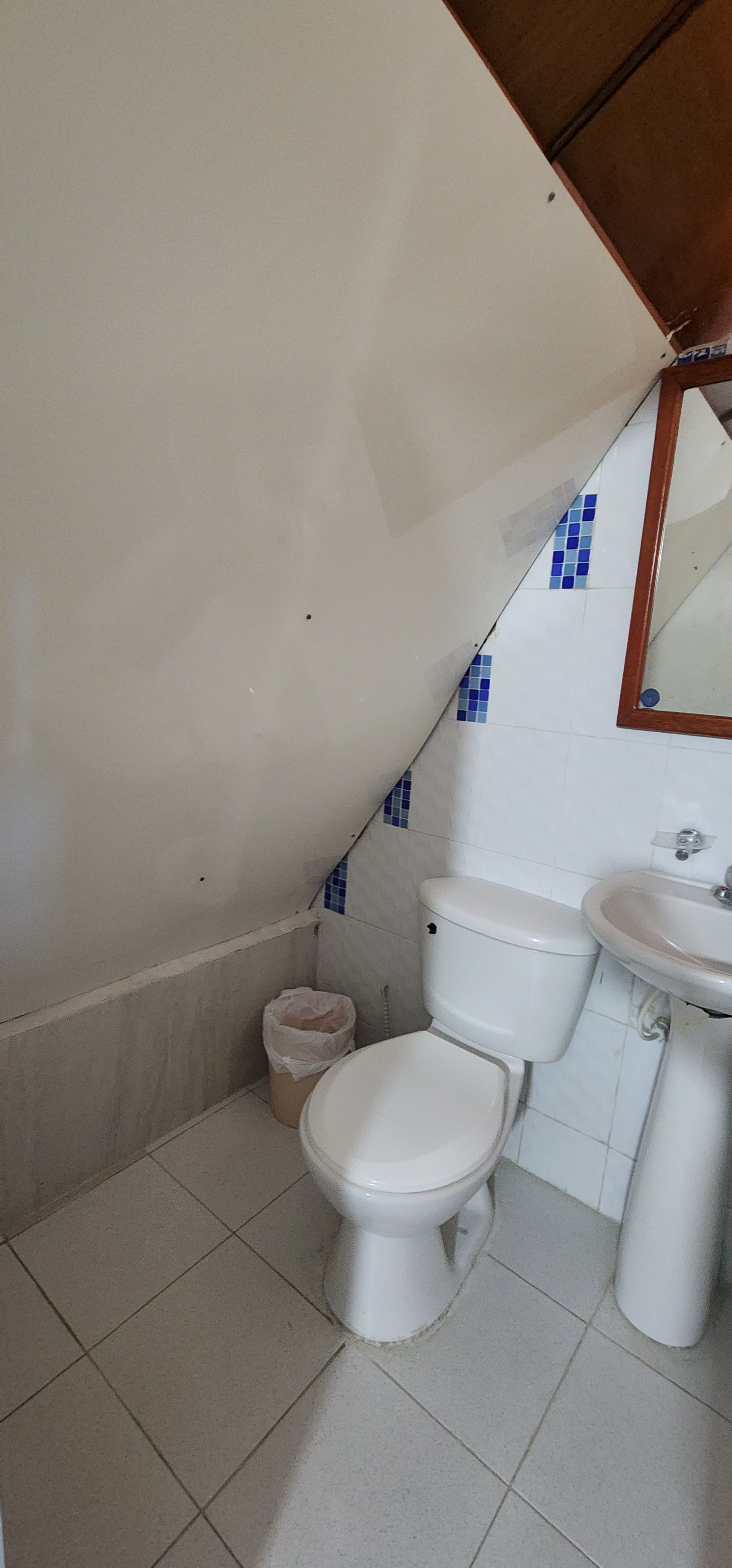 Un baño pequeño con techo inclinado, paredes alicatadas, inodoro blanco, lavabo con pedestal y espejo.
