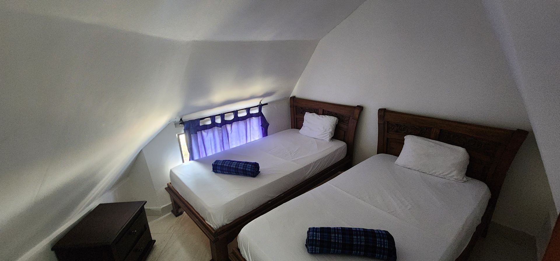 Un dormitorio con dos camas individuales de madera, ropa de cama blanca con toallas azules y una ventana con una cortina morada bajo una viga inclinada.
