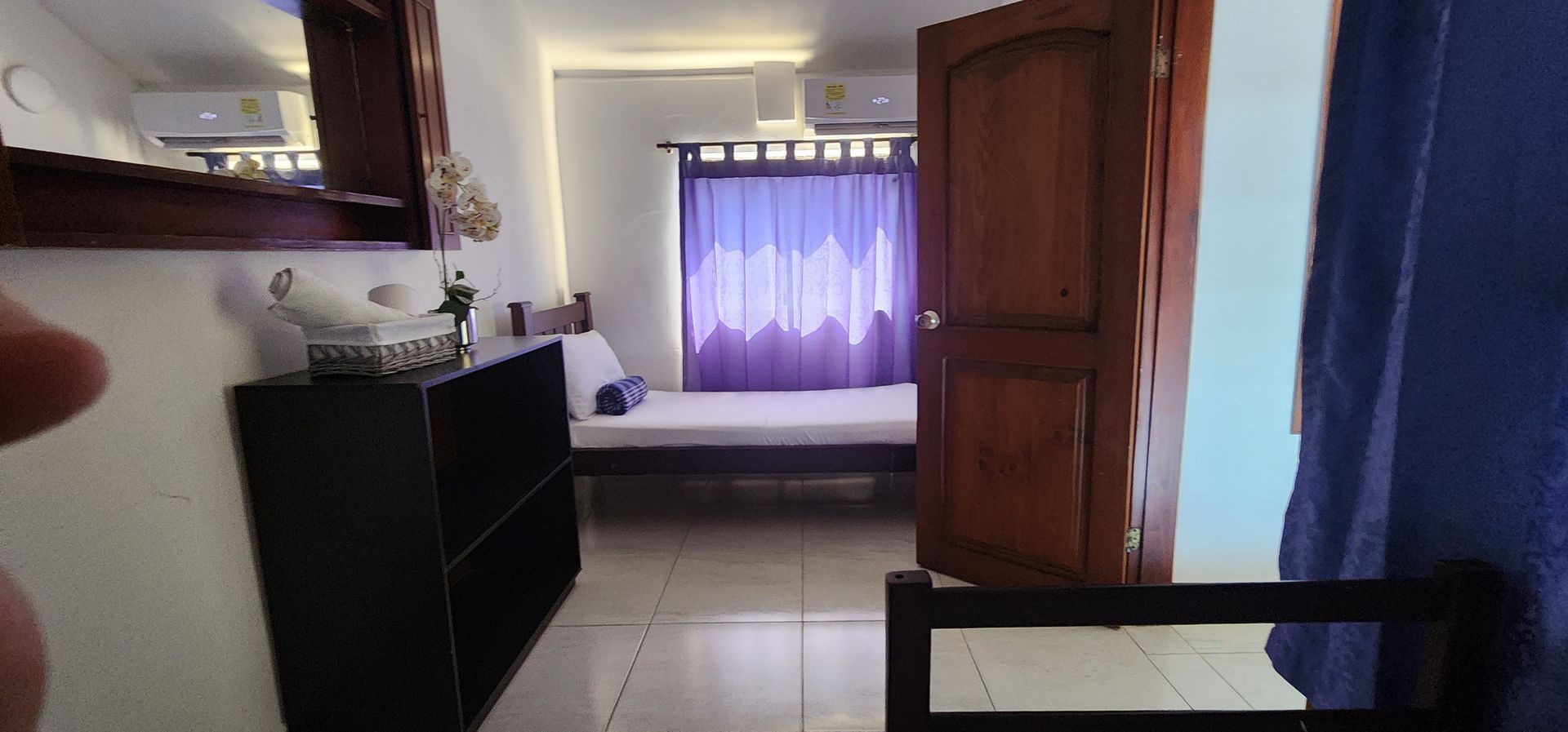 Una habitación con paredes blancas, una estantería negra y una puerta de madera abierta que da a un espacio con una cama y cortinas moradas.