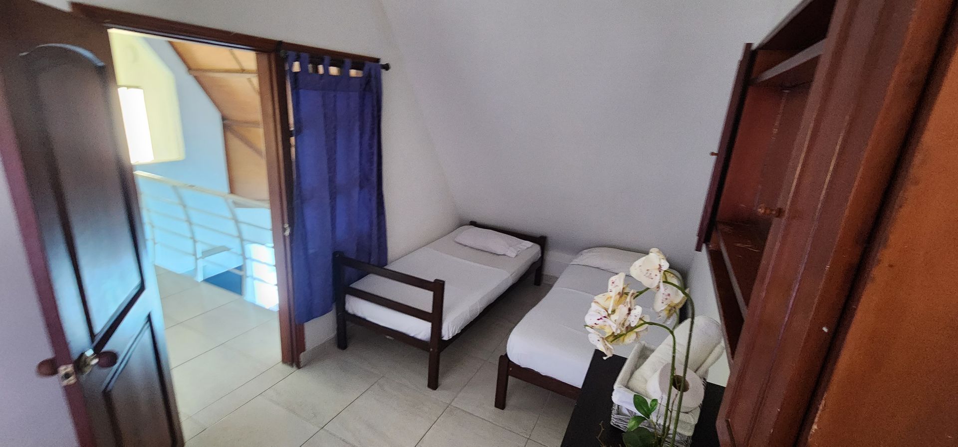 Un dormitorio con dos camas individuales, paredes blancas y un mueble de madera, visto a través de una puerta abierta con cortinas azules.