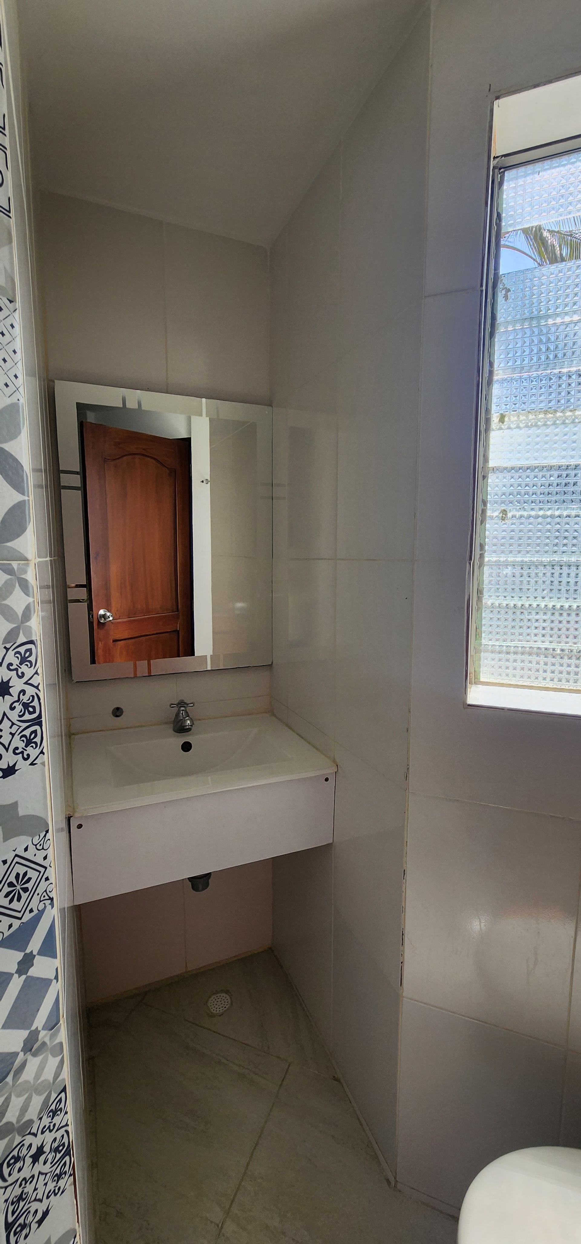 Un área de baño con lavabo, tocador suspendido, un espejo que refleja una puerta de madera y una ventana con azulejos a la derecha.