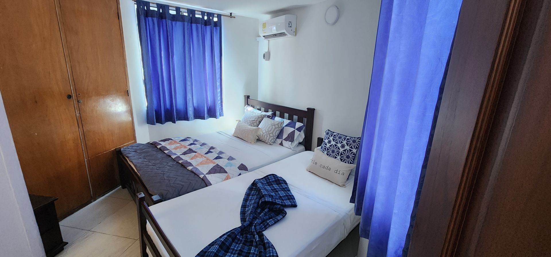 Una habitación de hotel con dos camas con sábanas blancas, detalles decorativos azules, cortinas azules y un armario de madera.