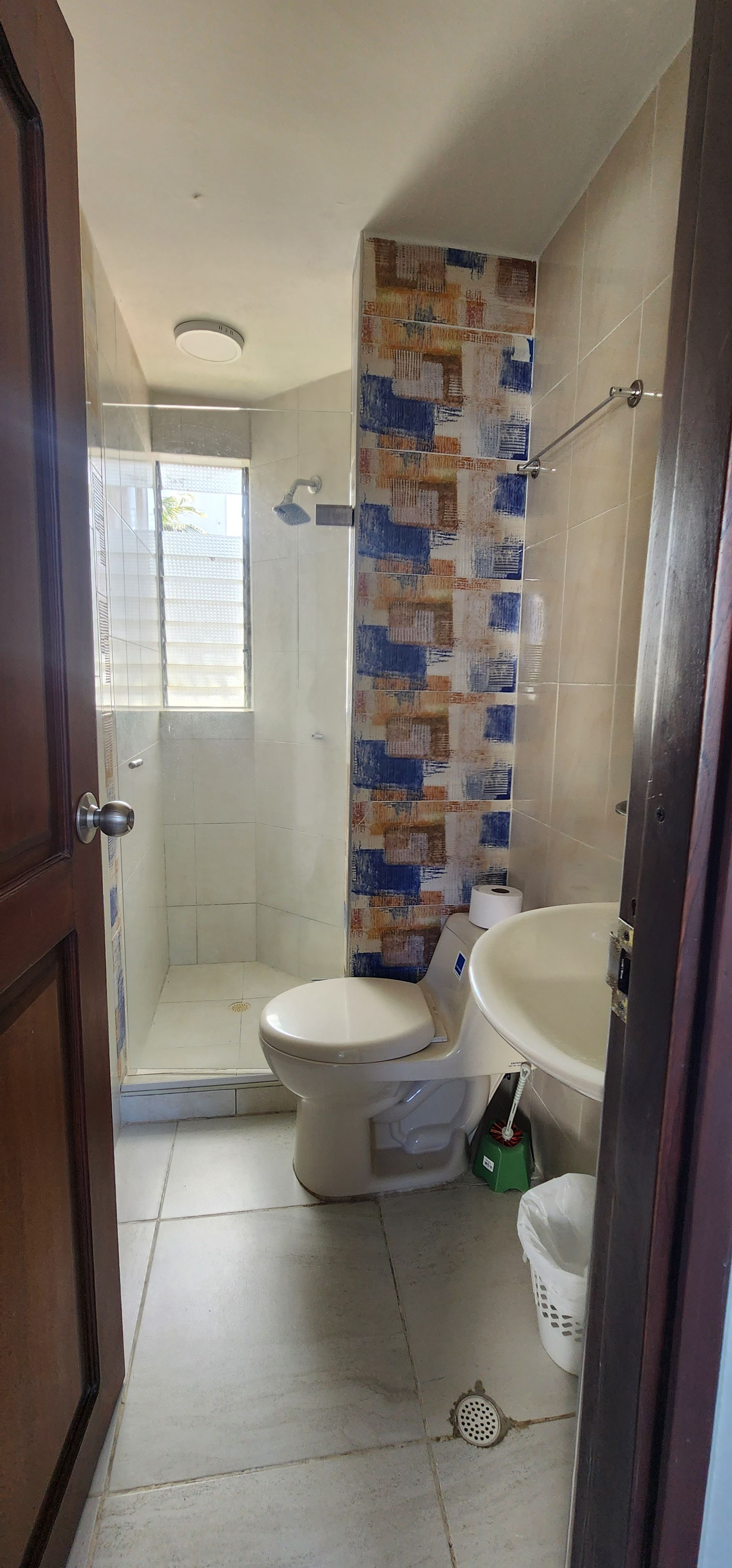 Un baño pequeño con suelo de baldosas, inodoro, lavabo, cabina de ducha en esquina y una pared decorativa con un estampado azul y beige.