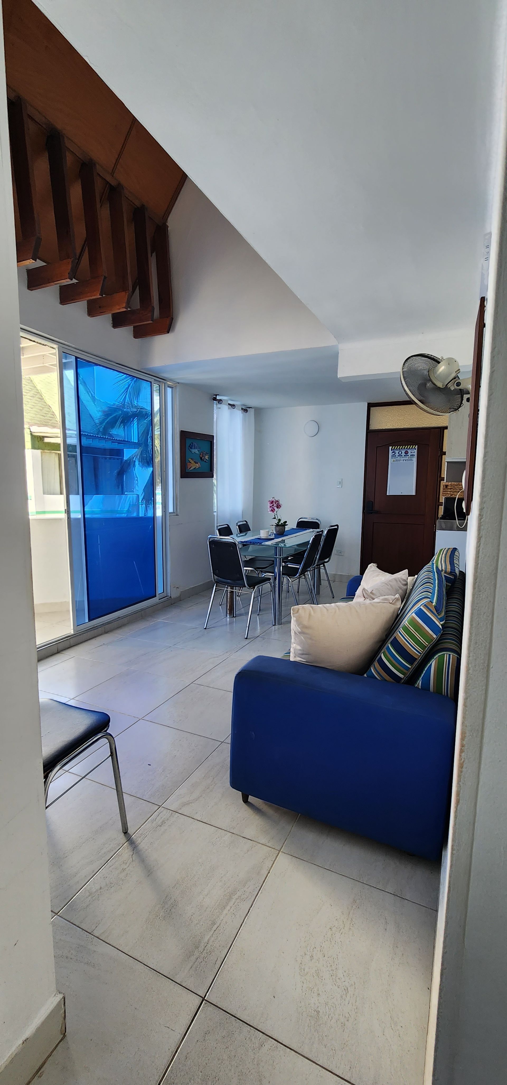 Una luminosa sala de estar cuenta con un sofá azul, un juego de comedor y puertas de cristal con cortinas azules, visible desde un pasillo blanco.