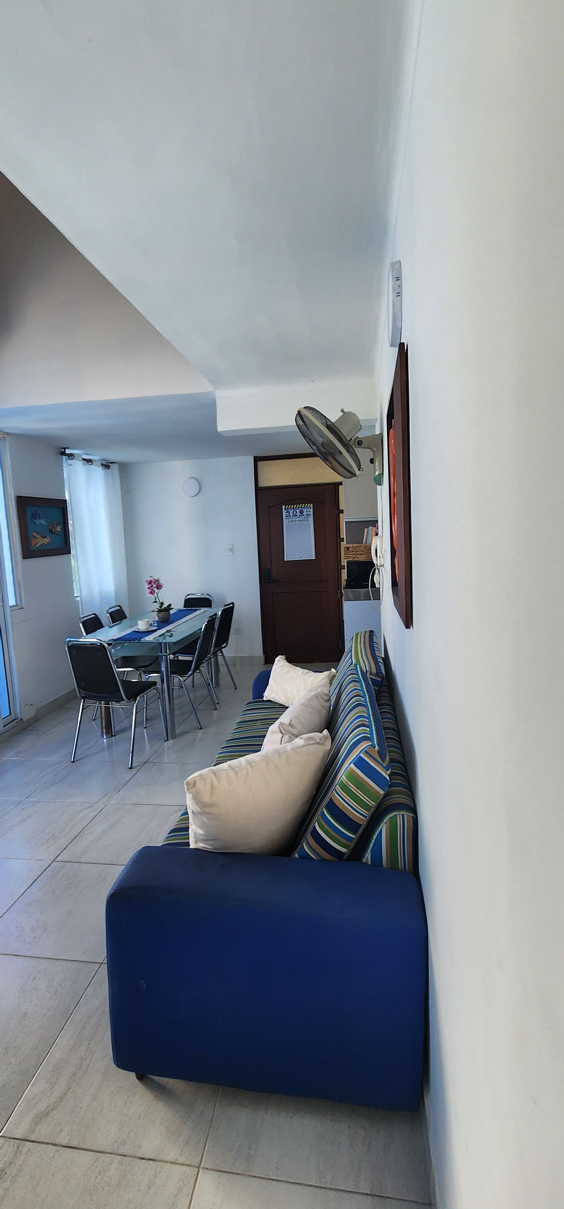 En una sala de estar hay un sofá azul con cojines estampados, con una mesa de comedor y sillas al fondo.