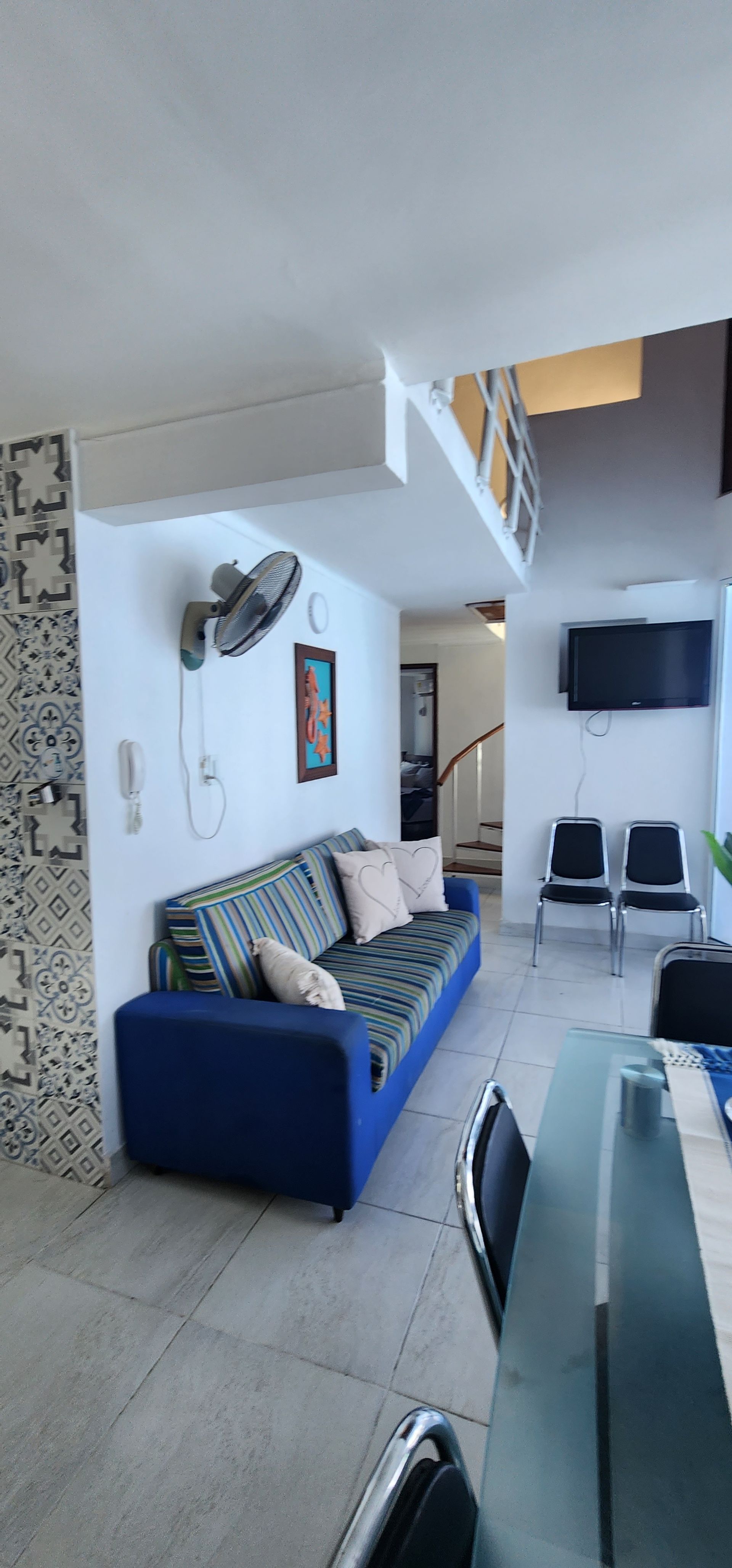 Vista interior de una habitación con un sofá azul, un ventilador de techo, cuadros, sillas de comedor y un televisor montado en la pared.