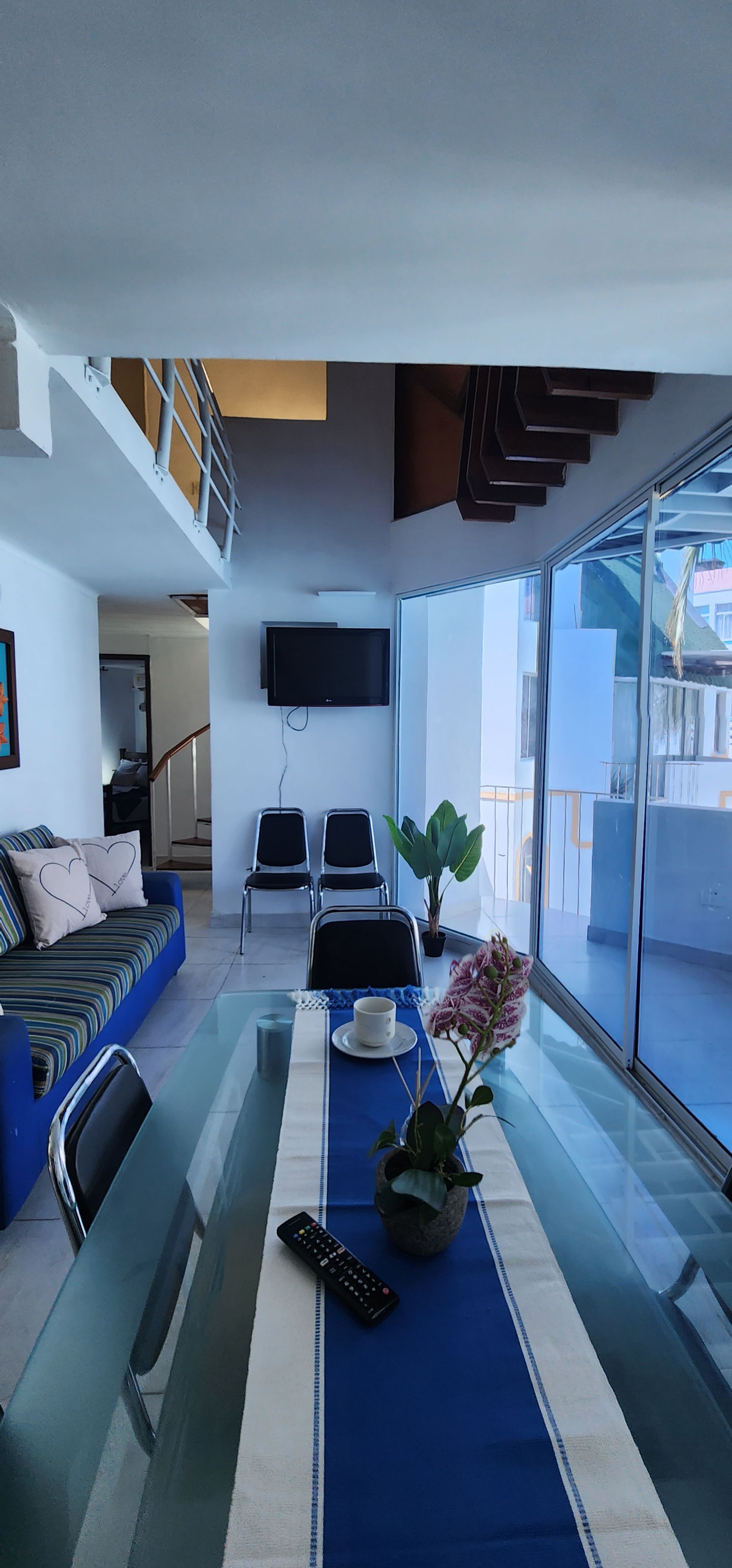Una larga mesa de comedor con un camino de mesa azul se encuentra en una habitación que cuenta con un sofá azul, un televisor montado en la pared y una escalera de caracol.