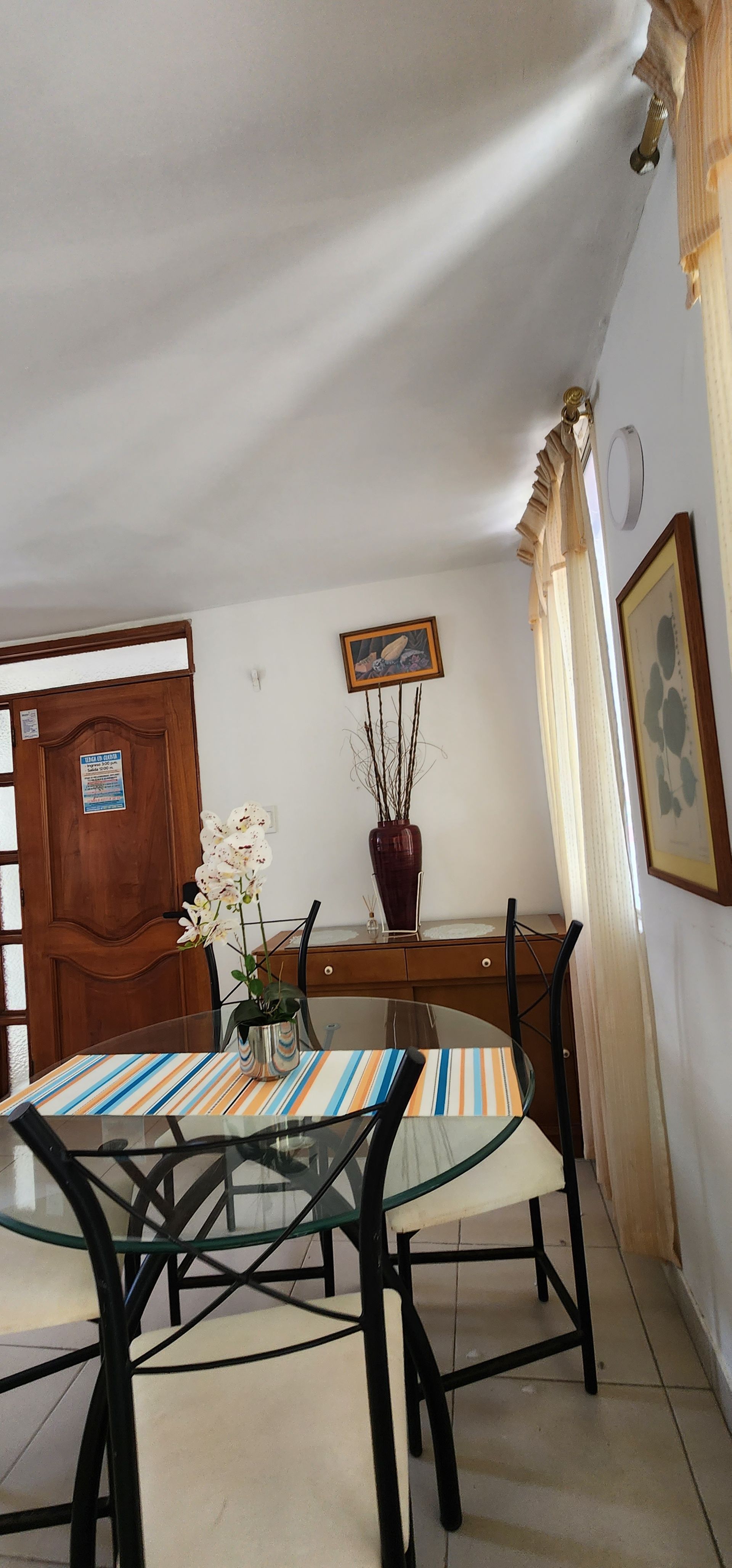 Un comedor con mesa y sillas con tablero de cristal, una mesa consola de madera, un jarrón con flores y cuadros enmarcados en la pared.