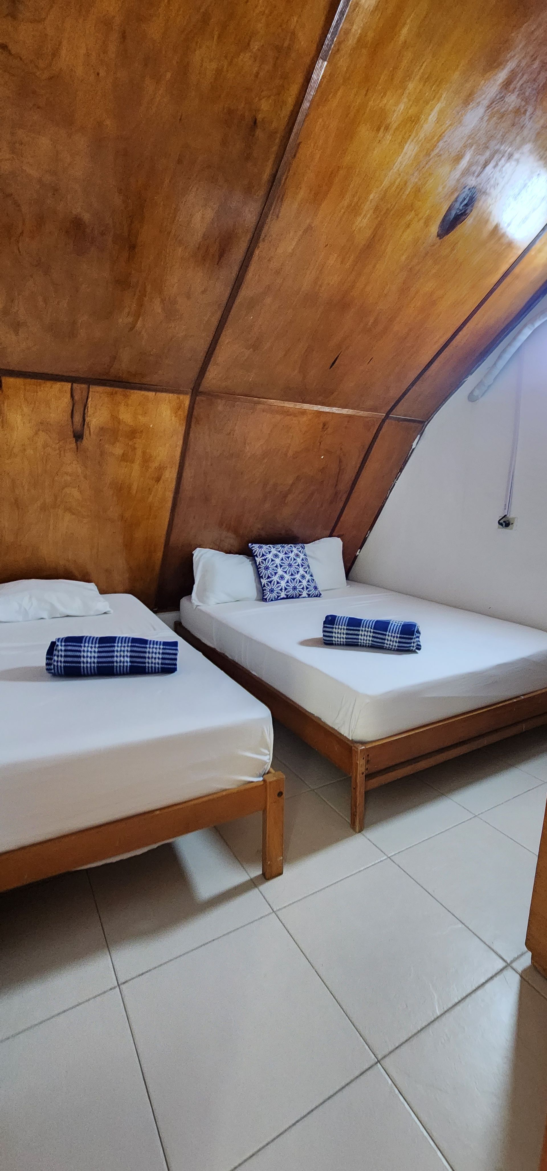 Dos camas con sábanas blancas y detalles estampados en azul en una habitación con un singular techo inclinado revestido de paneles de madera.