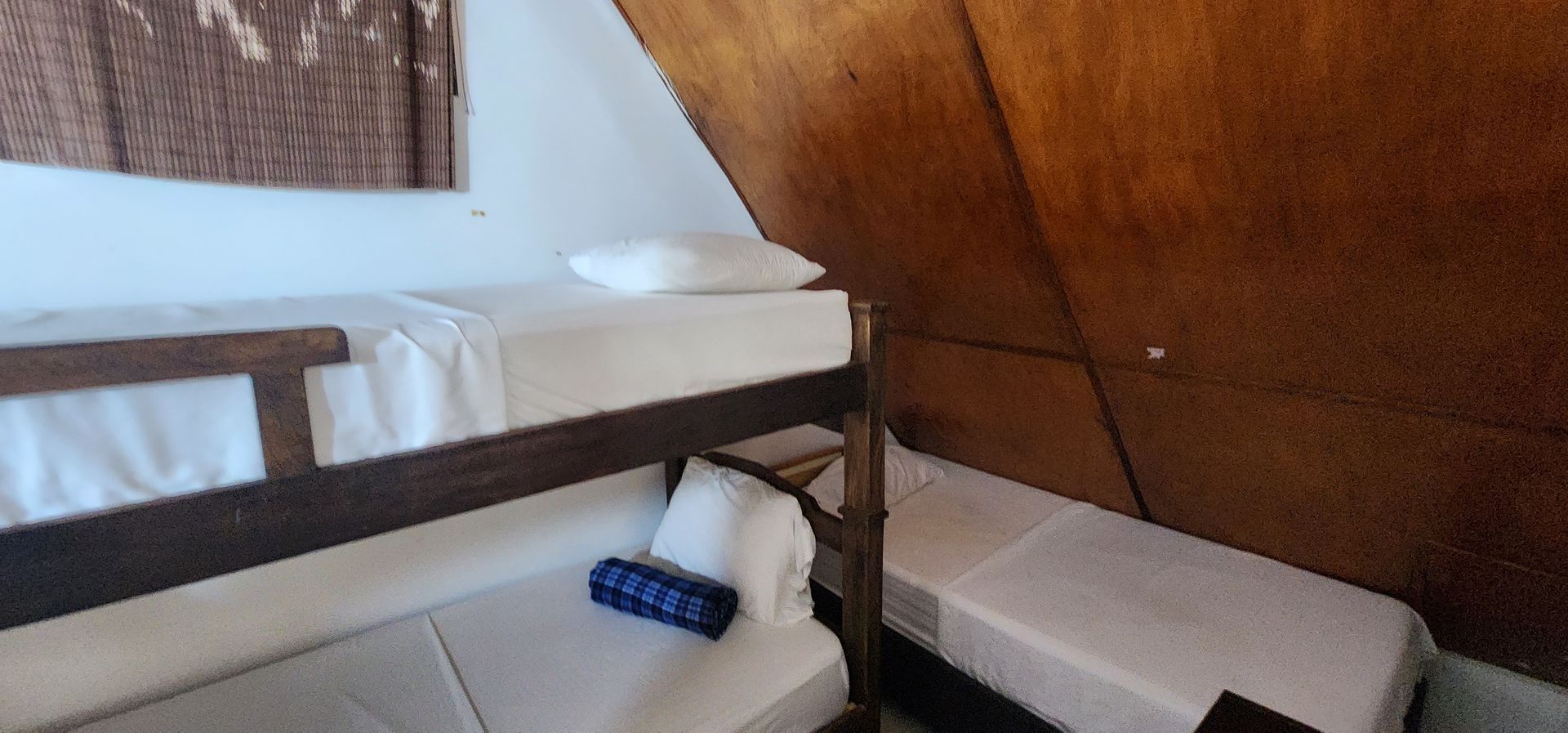 Una litera de madera y una cama individual contigua con sábanas y almohadas blancas en una habitación con paredes inclinadas y texturizadas.