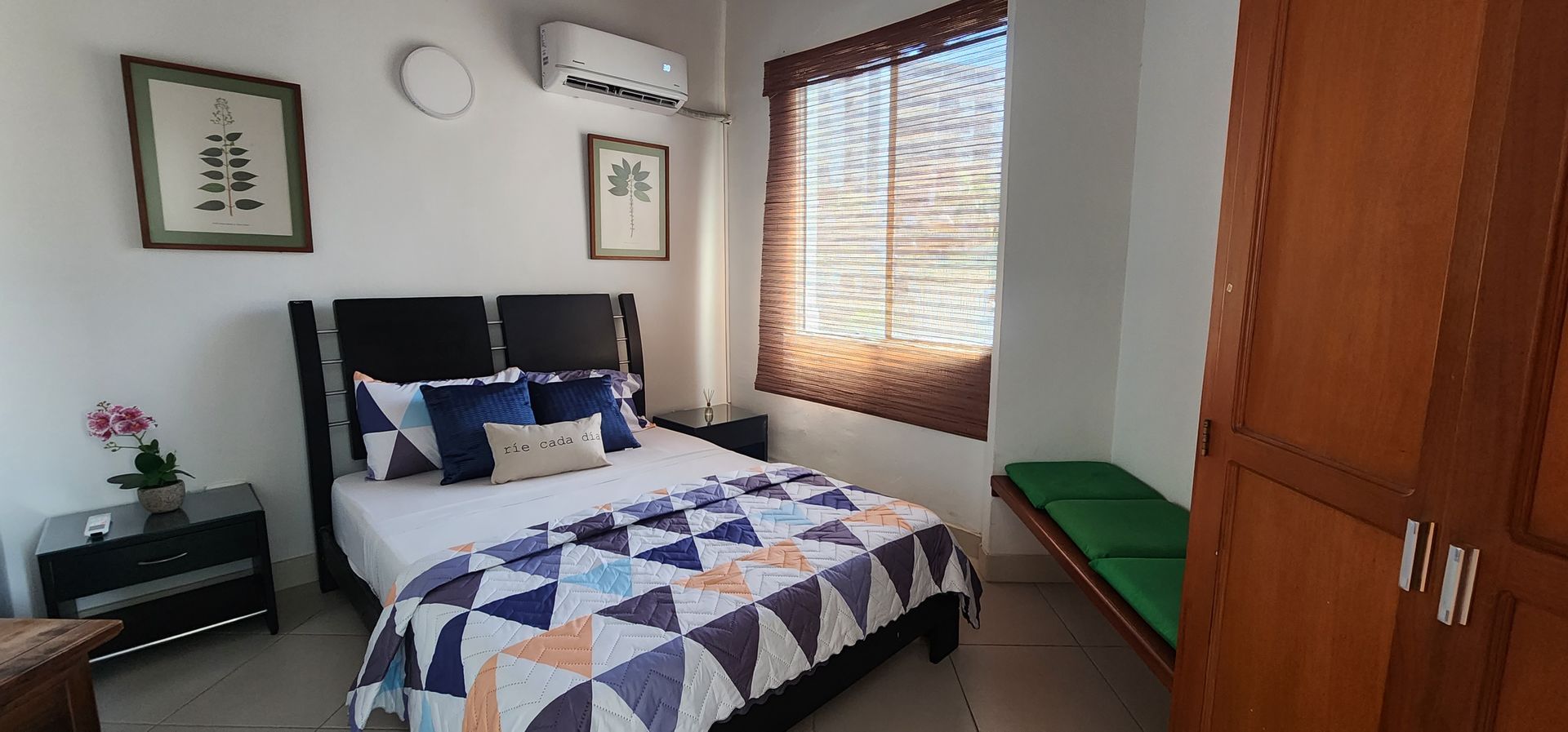 Un dormitorio con una cama con una colcha de estampado geométrico, dos láminas botánicas enmarcadas, un armario de madera y un banco.