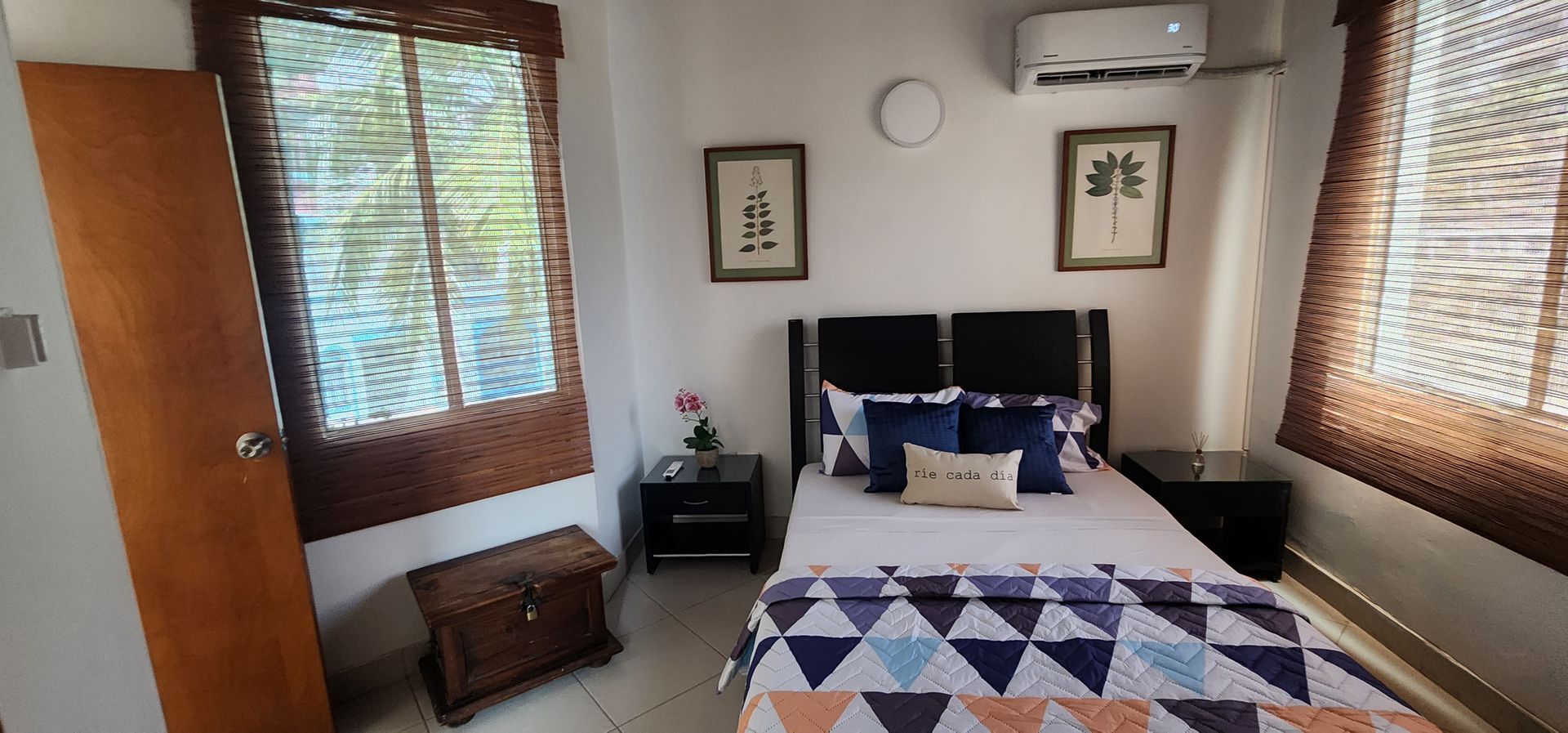 Un dormitorio luminoso con una cama de estampado geométrico, dos mesitas de noche pequeñas, cuadros enmarcados en la pared y persianas de bambú.