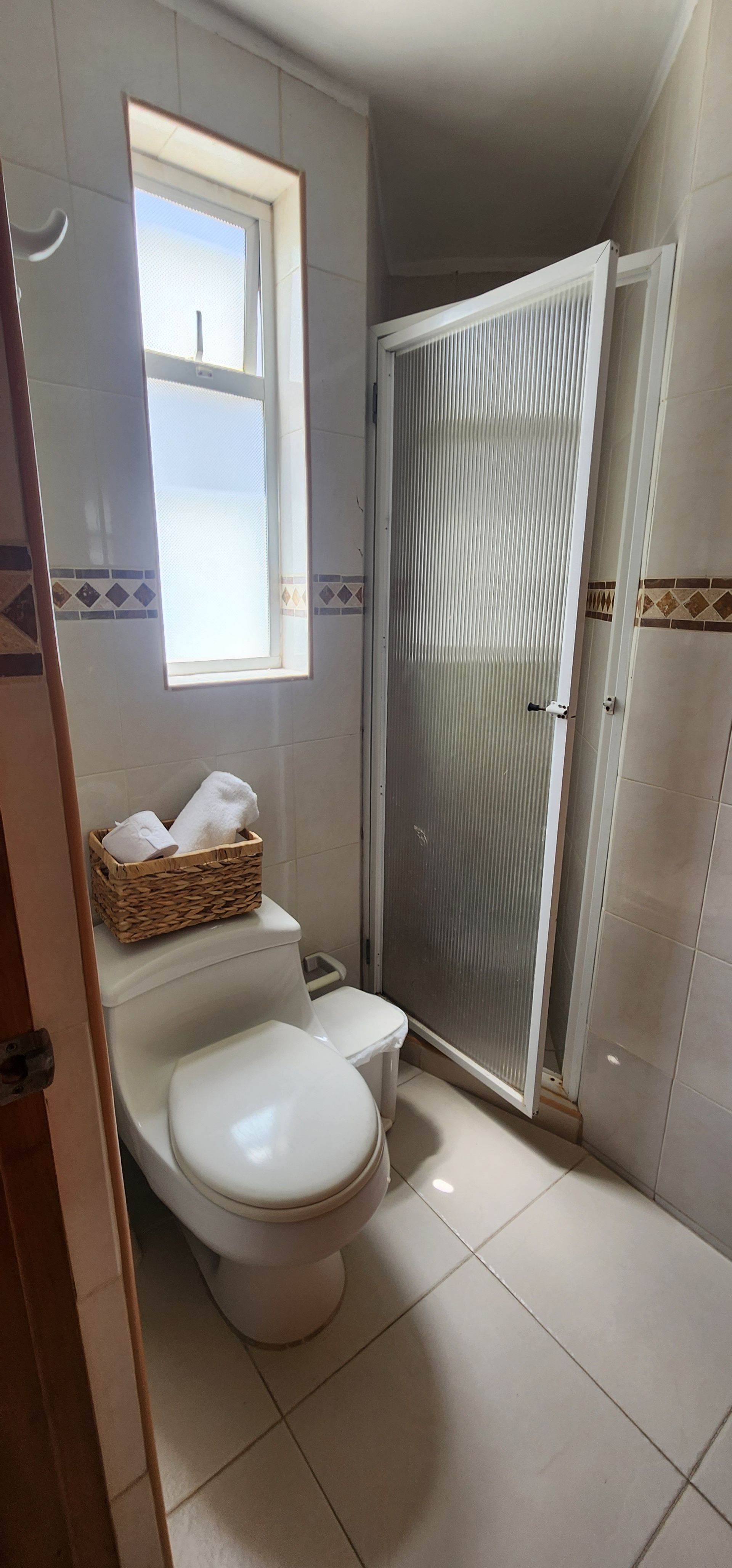 Un baño con paredes de azulejos de color claro, un inodoro con una cesta para toallas encima y una puerta de ducha con paneles de vidrio.