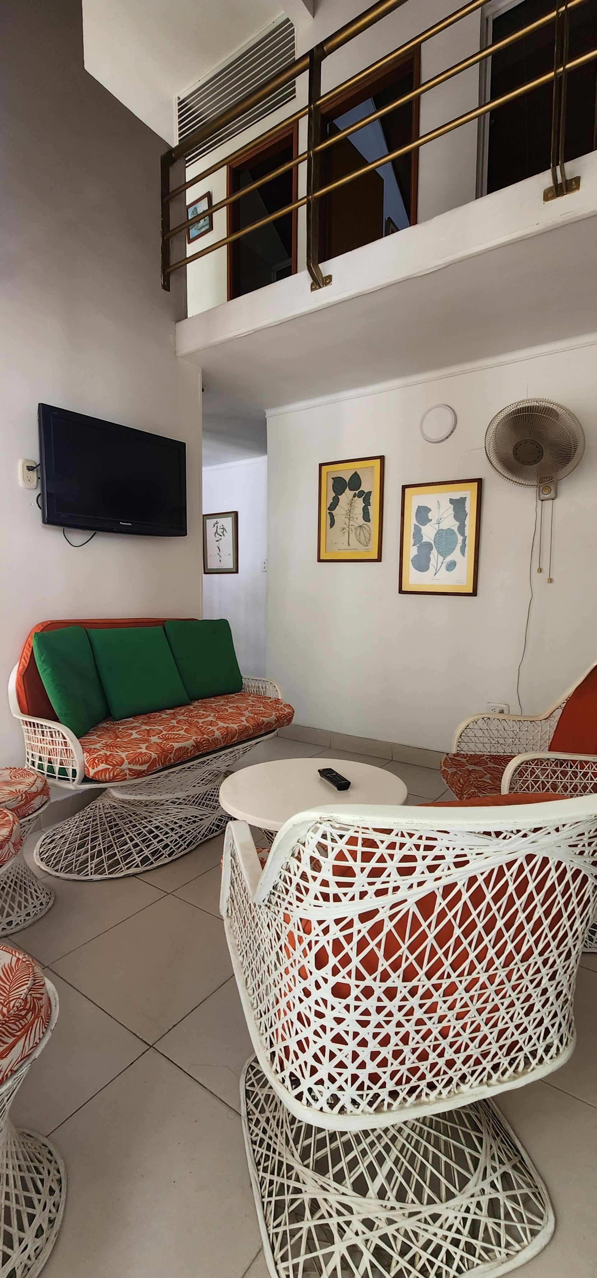 Una sala de estar con un sofá de cojines verdes, sillas blancas estampadas, una pequeña mesa redonda y obras de arte en la pared, bajo un balcón tipo loft.