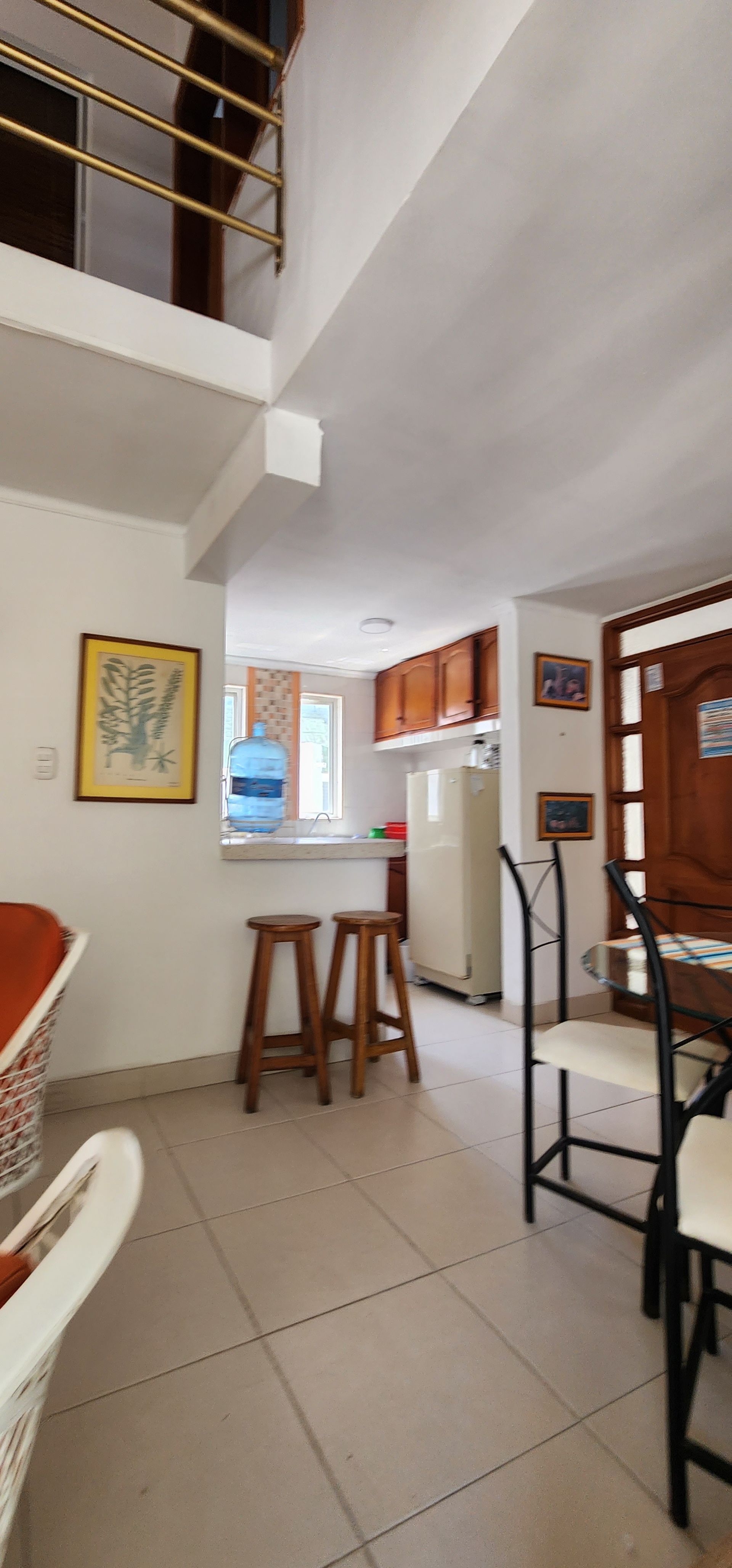 Vista interior que muestra una zona de cocina abierta con una encimera, dos taburetes de madera, suelo de baldosas de color claro y una escalera.