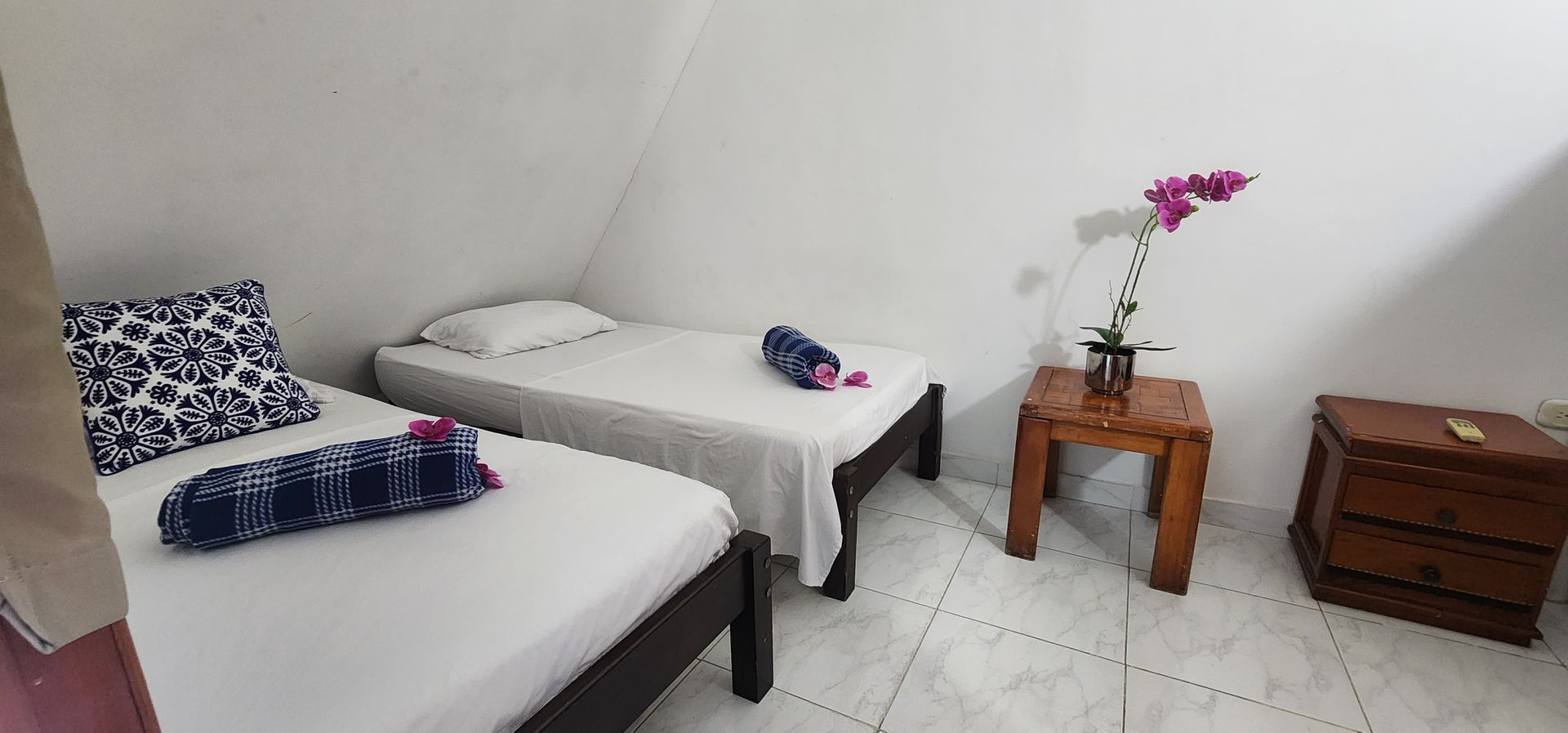 Un dormitorio minimalista con dos camas individuales con almohadas estampadas, una pequeña mesita de noche de madera con flores y una cómoda.