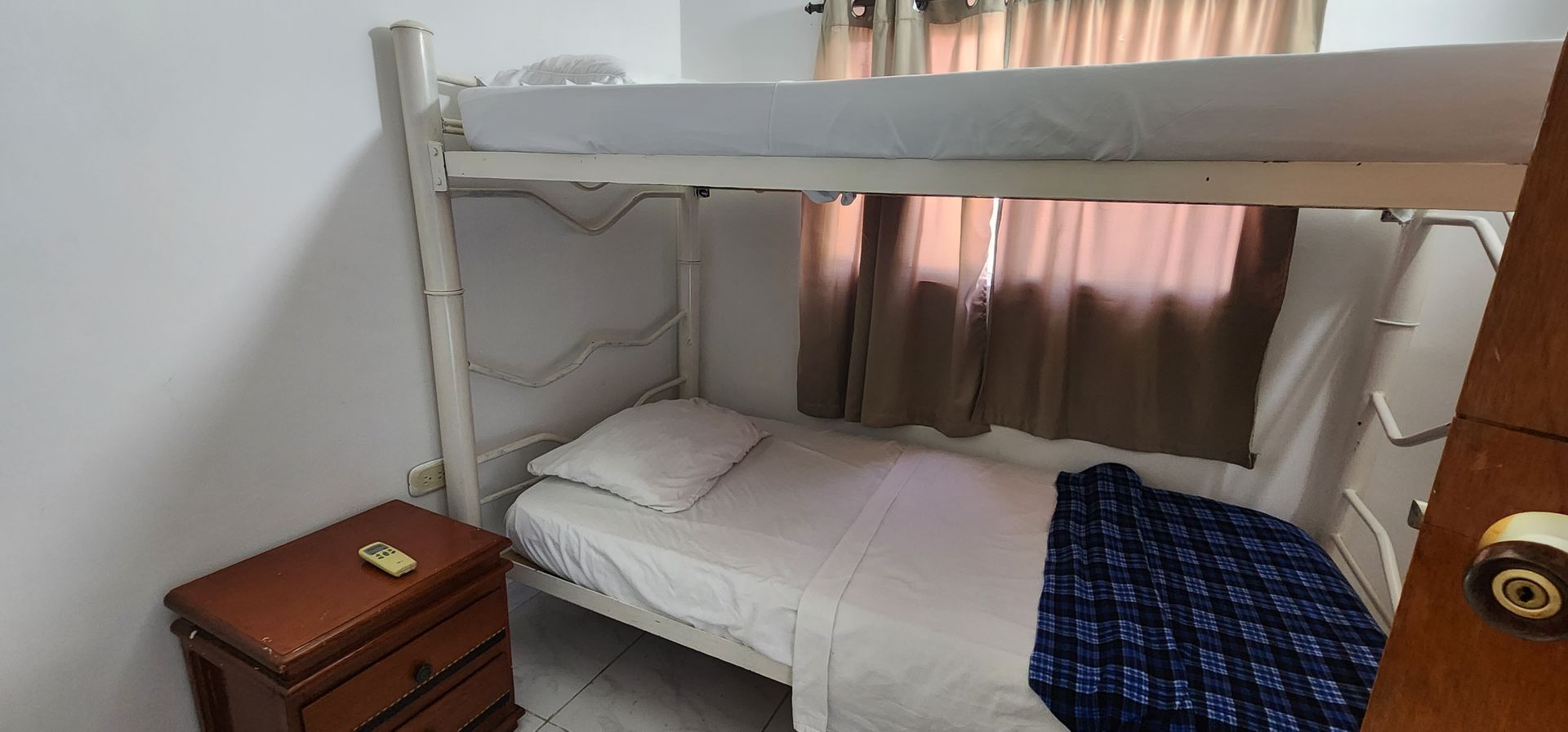 Una litera con estructura y ropa de cama blancas, una pequeña mesita de noche de madera y cortinas marrones en un dormitorio compacto.