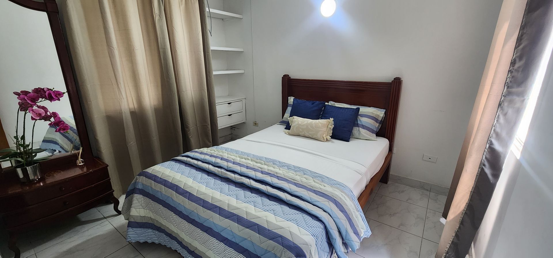 Un dormitorio acogedor con una cama de madera con ropa de cama a rayas azules, una cómoda a juego con espejo y estantes blancos.
